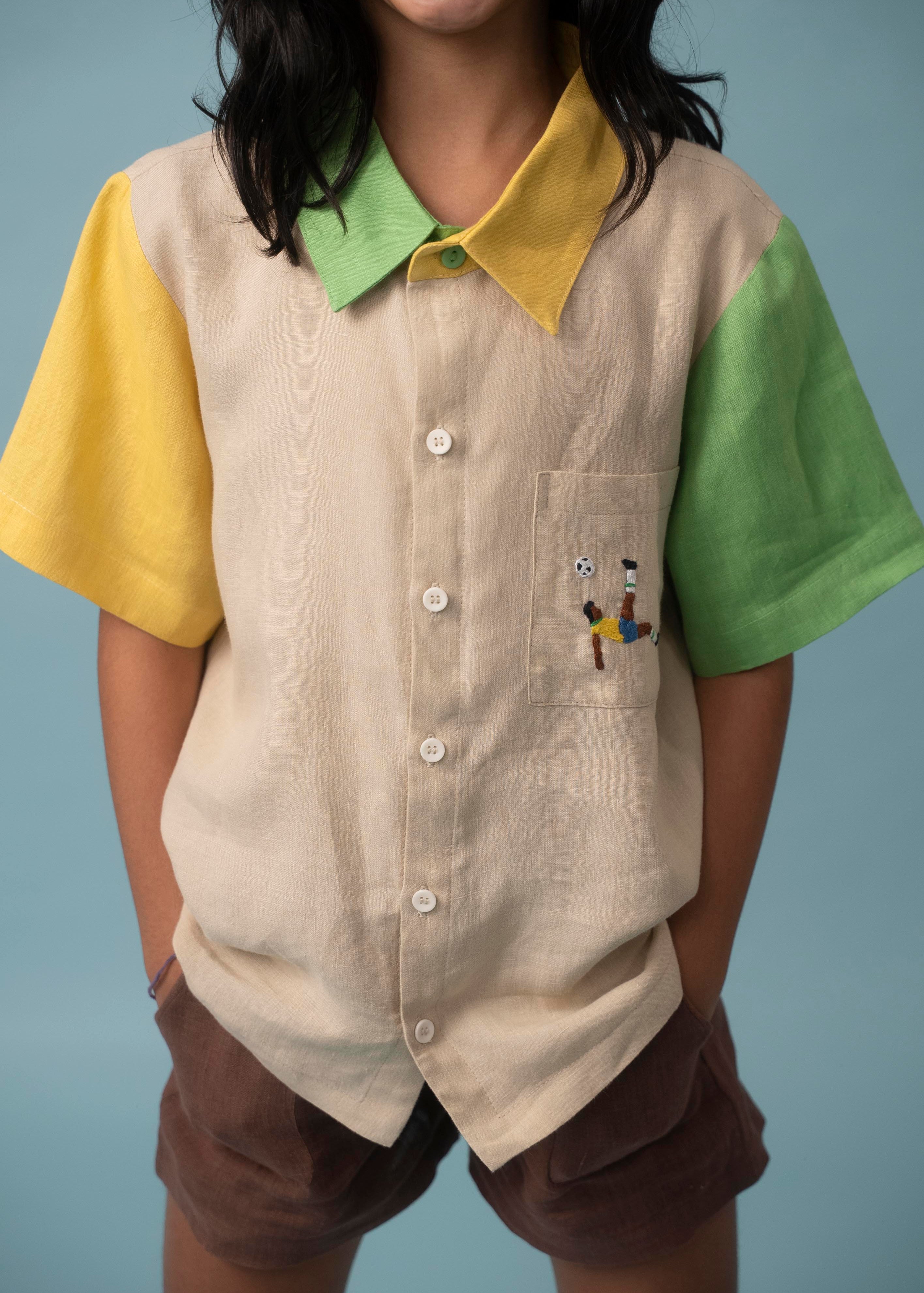 Menina usando camisa infantil de linho bege, verde e amarelo com bordado de jogador de futebol no bolso