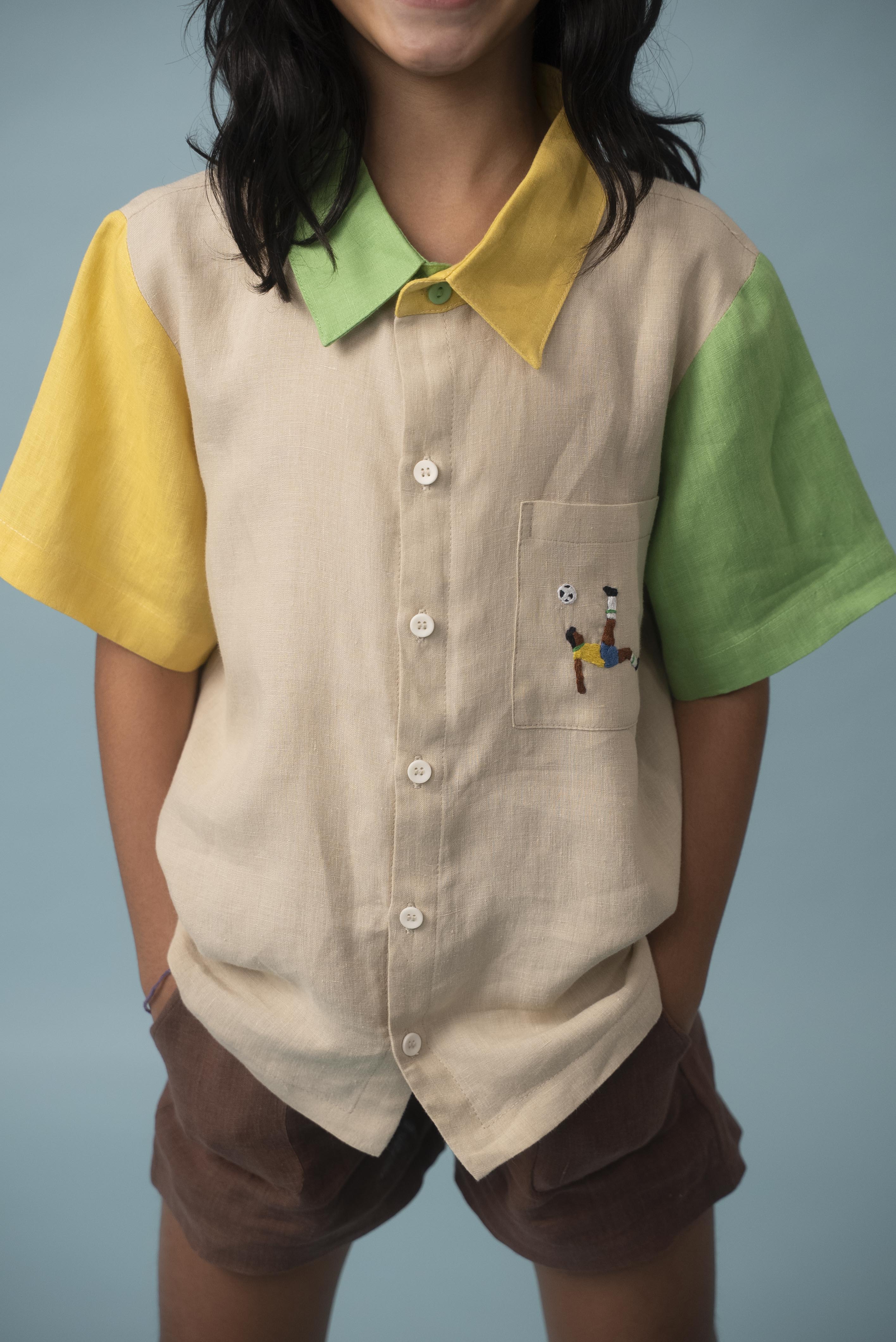 Menina usando camisa infantil de linho bege, verde e amarelo com bordado de jogador de futebol no bolso