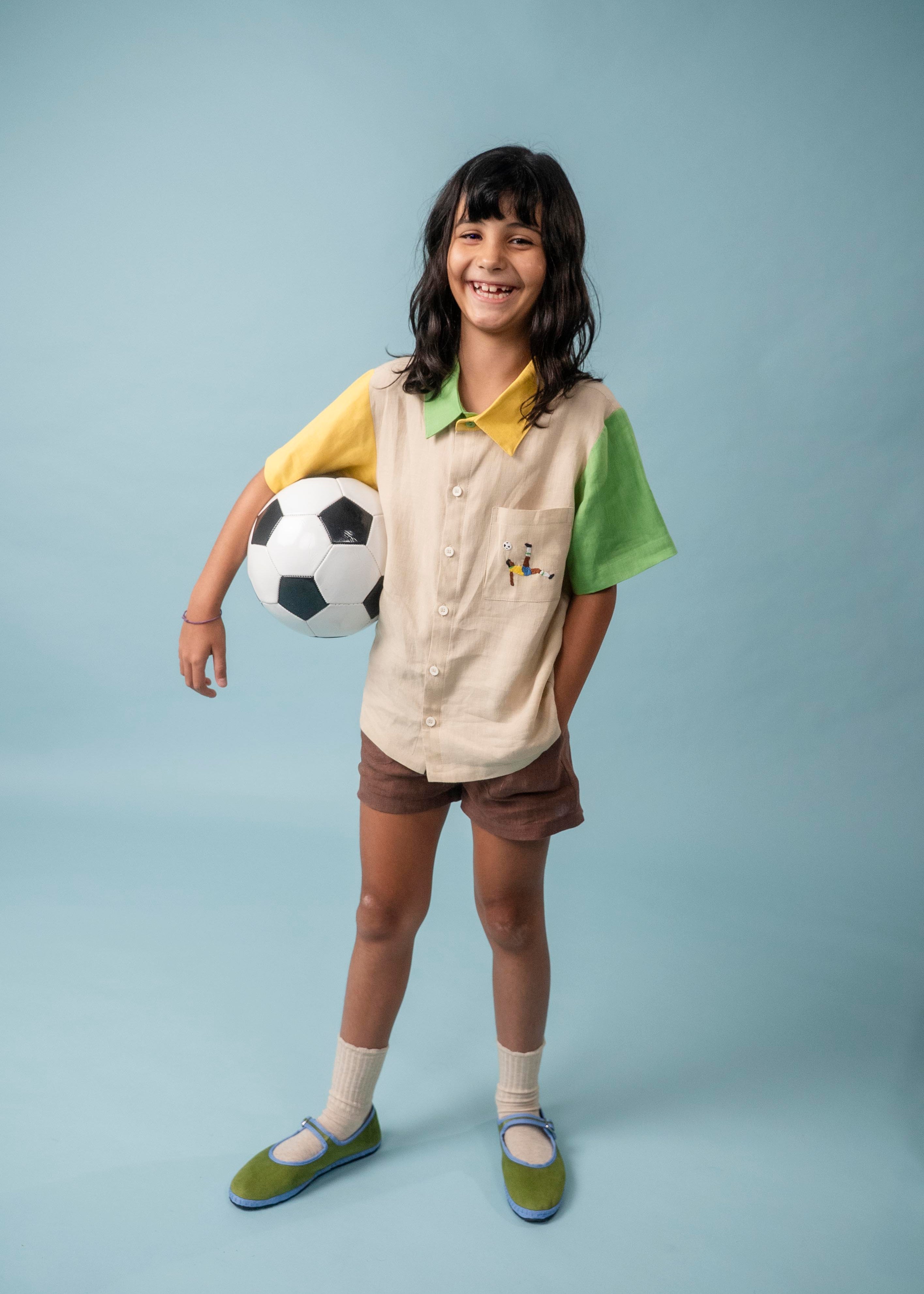 Menina usando camisa infantil em linho puro com bordado de futebol, combinada com shorts marrom e bola