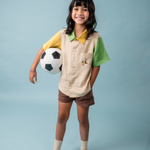 Menina usando camisa infantil de linho bege, verde e amarelo com bordado de jogador de futebol no bolso, combinando com shorts marrom.