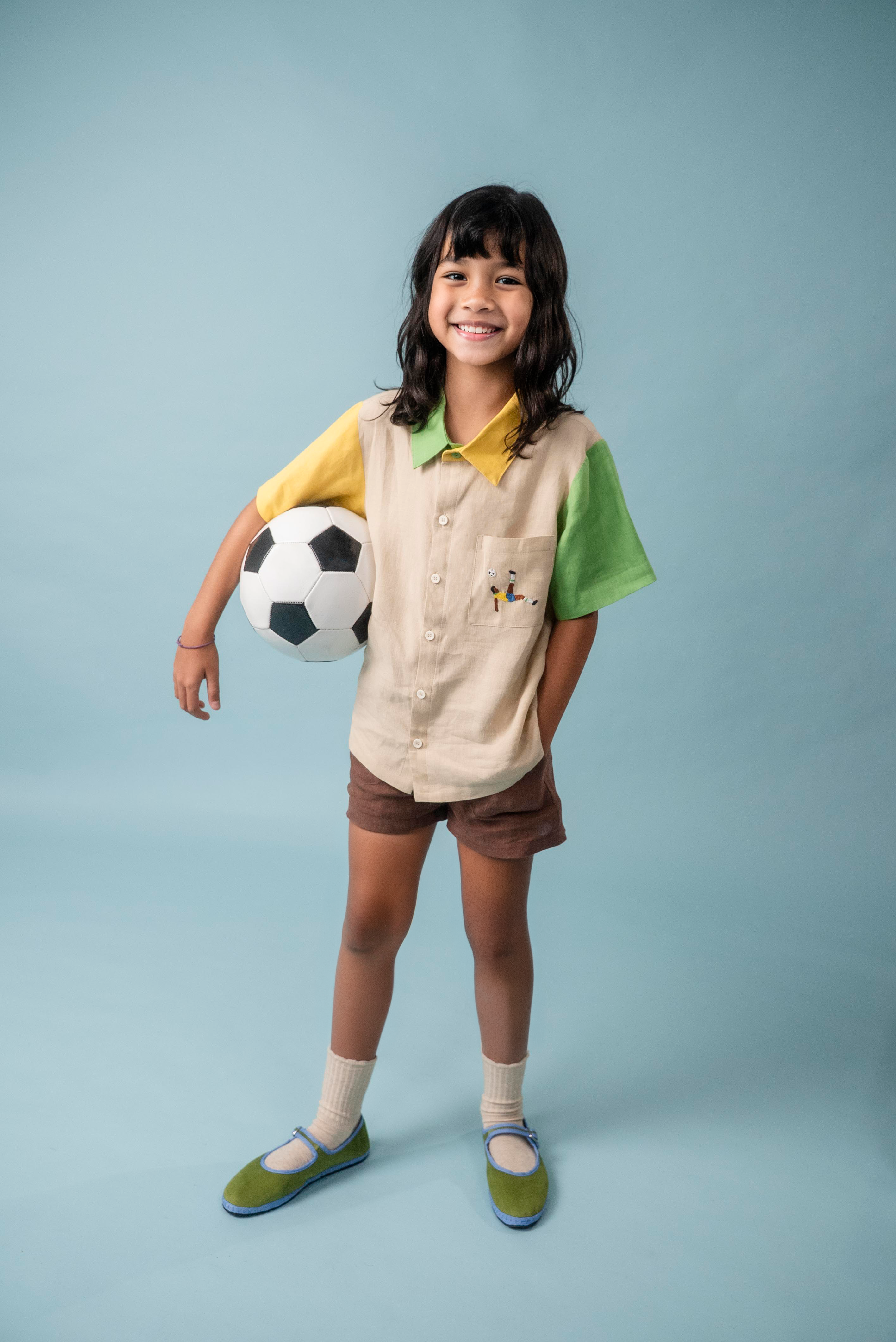 Menina usando camisa infantil de linho bege, verde e amarelo com bordado de jogador de futebol no bolso, combinando com shorts marrom.