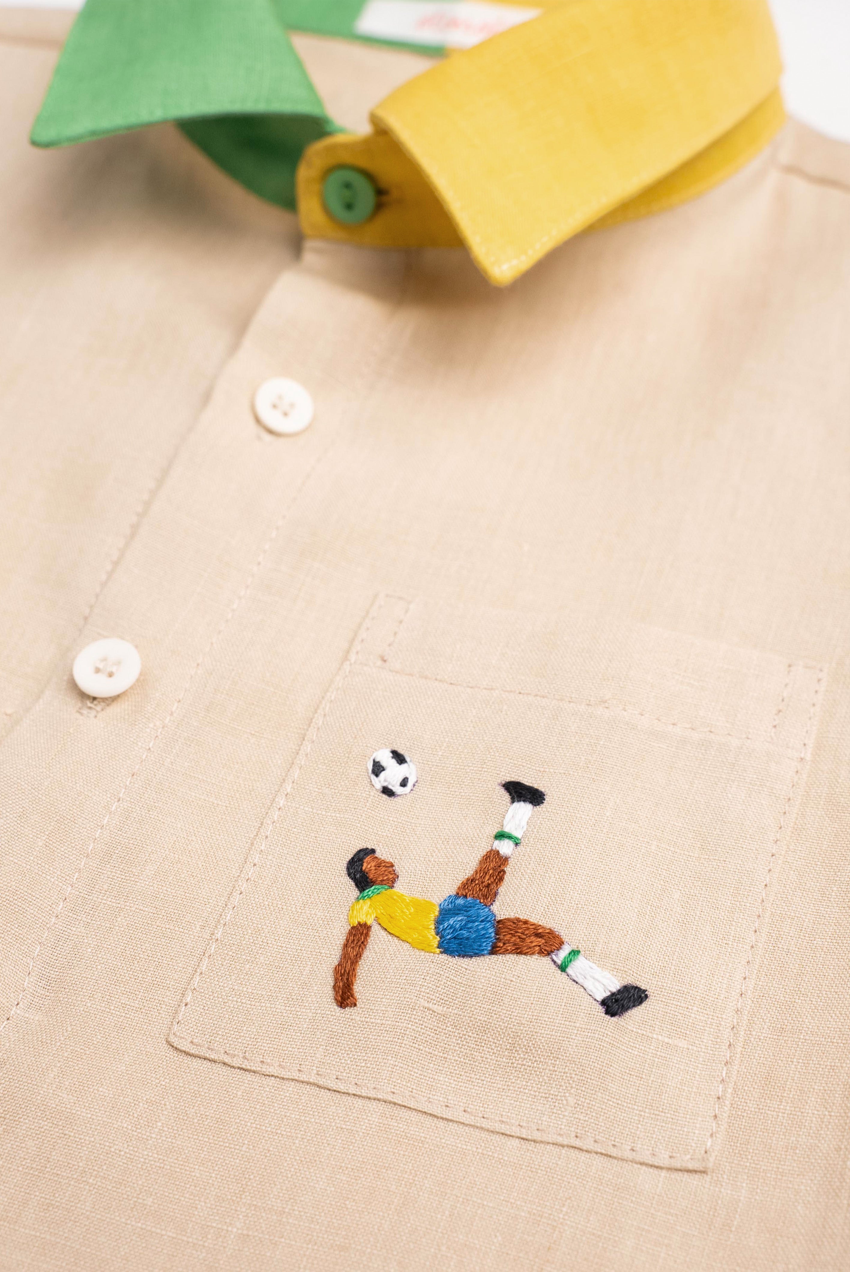 Detalhe do bordado à mão de jogador de futebol inspirado na bicicleta de Pelé em camisa infantil de linho puro