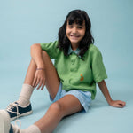 Menina usando camisa infantil de linho verde com bordado de cachorro caramelo, combinada com shorts azul claro