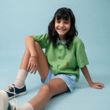 Menina usando camisa infantil de linho verde com bordado de cachorro caramelo, combinada com shorts azul claro