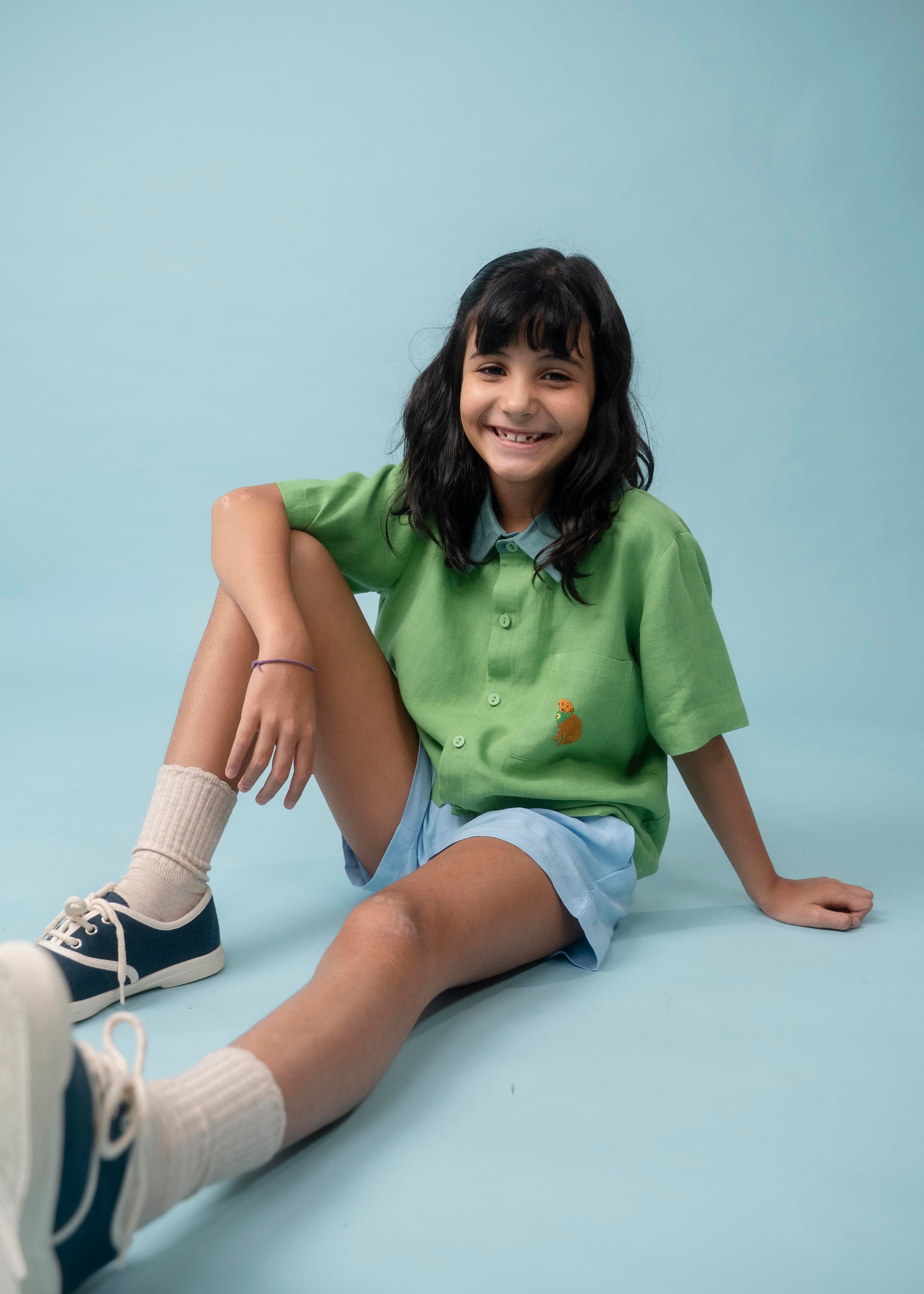 Menina usando camisa infantil de linho verde com bordado de cachorro caramelo, combinada com shorts azul claro