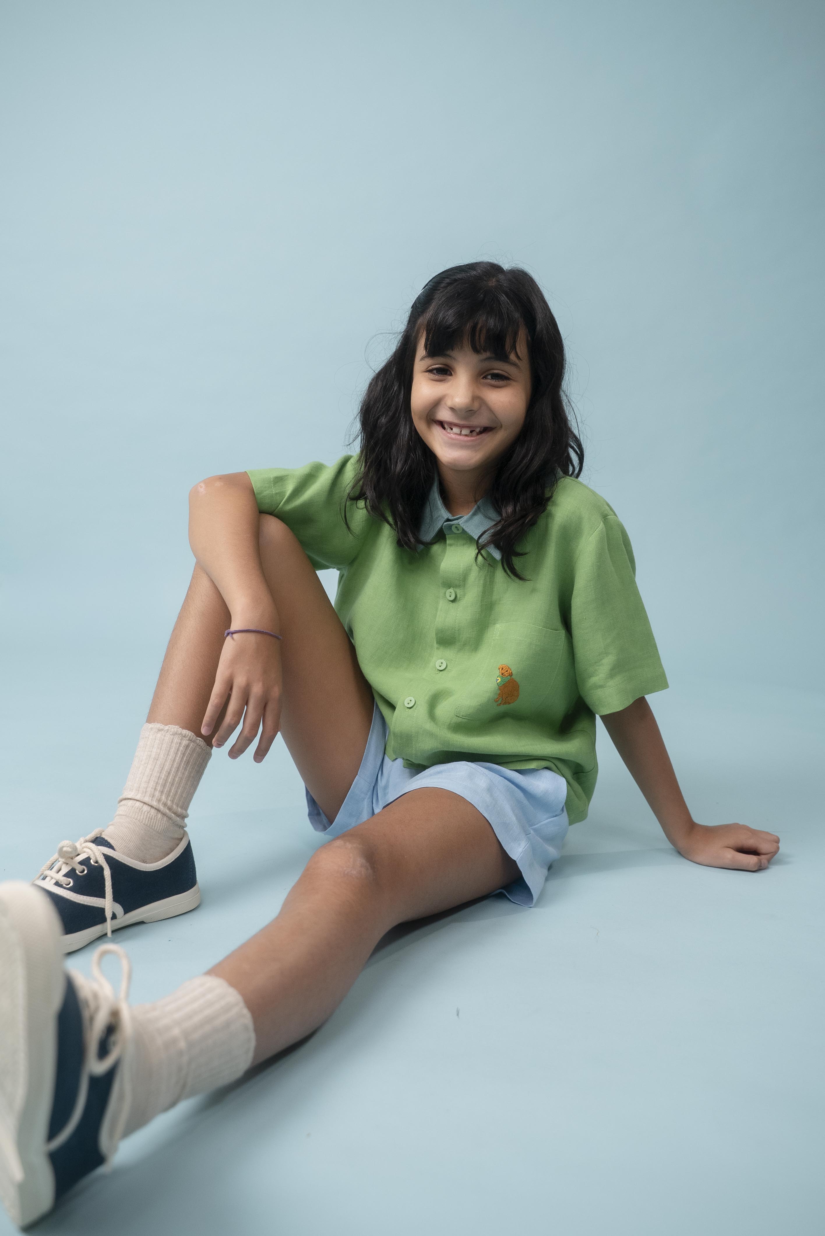 Menina usando camisa infantil de linho verde com bordado de cachorro caramelo, combinada com shorts azul claro
