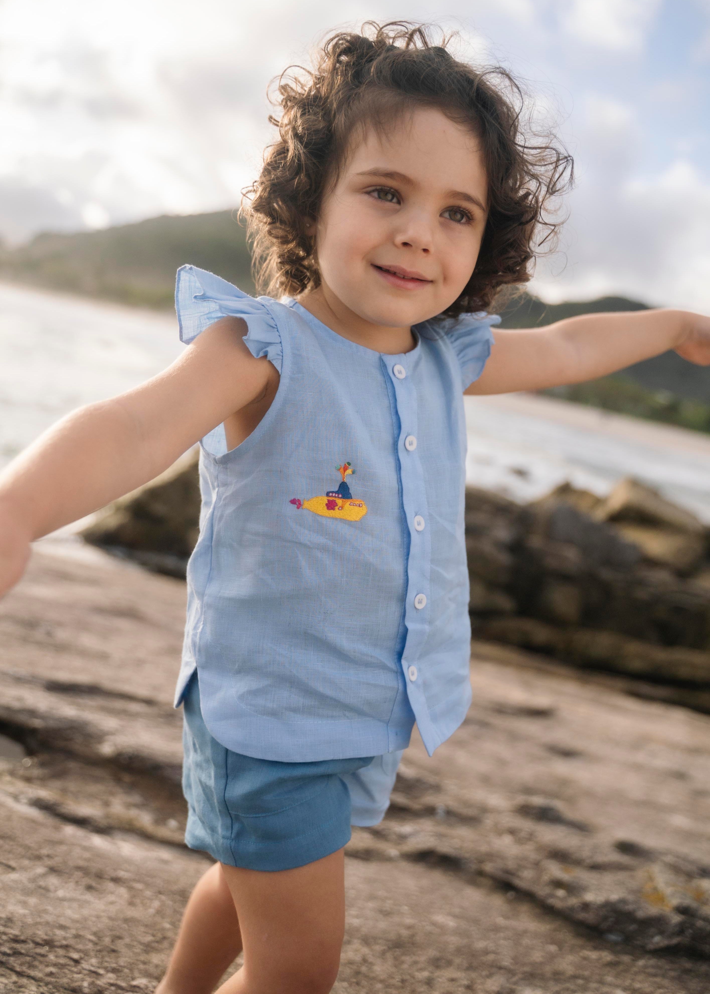 Menina vestindo camisa em linho azul com babado nas mangas, botões brancos frontais e bordado à mão de submarino amarelo inspirado em Yellow Submarine, dos Beatles.