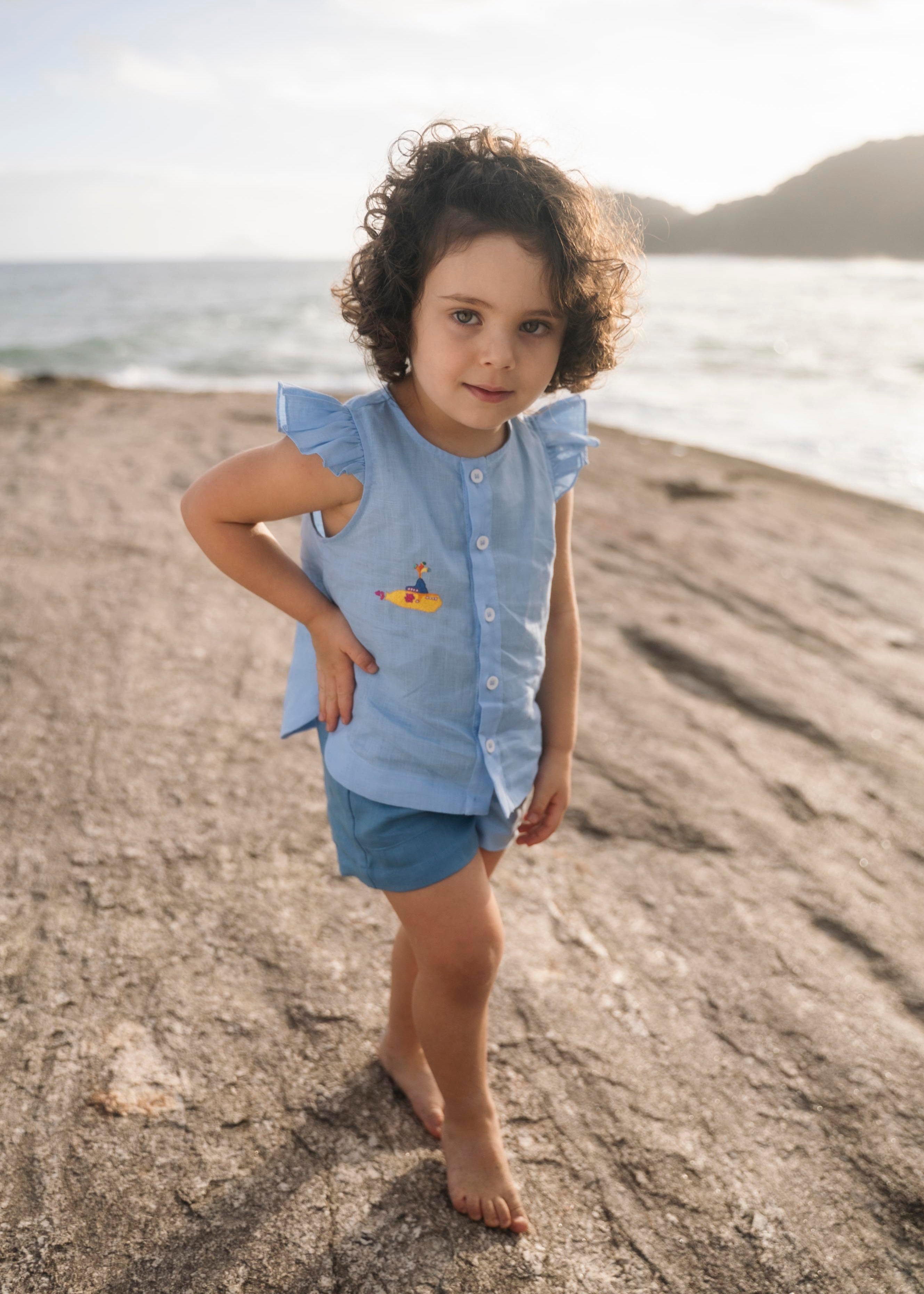 Menina vestindo camisa em linho azul com babado nas mangas e bordado à mão de submarino amarelo inspirado em Yellow Submarine, dos Beatles, com shorts combinando.