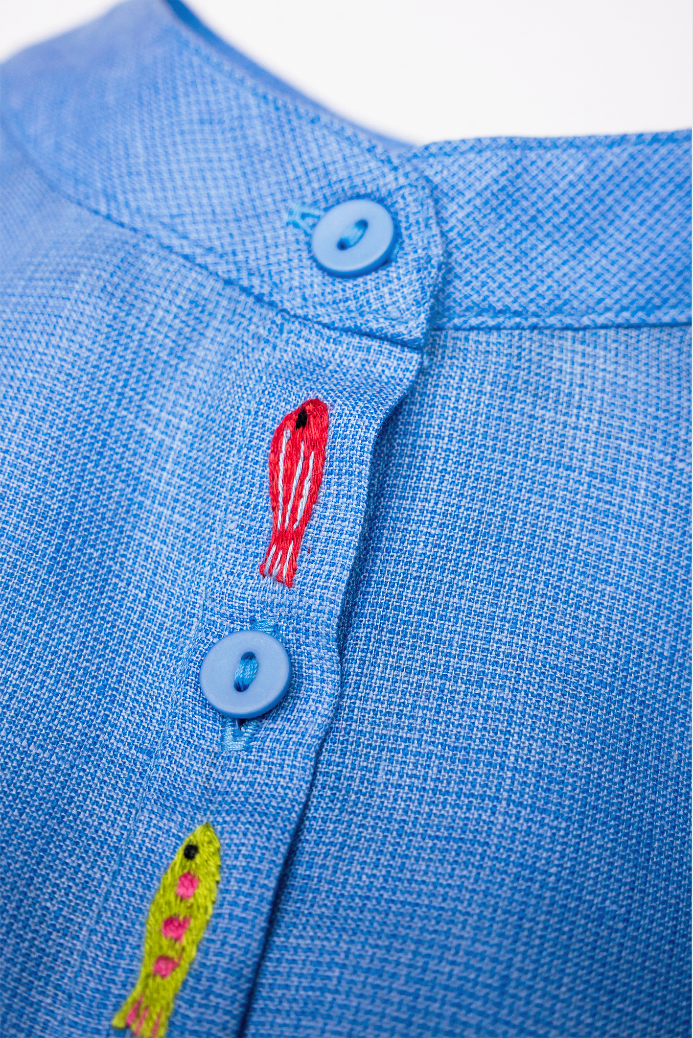 Close dos peixinhos bordados à mão entre os botões da Camisa Bento em linho azul fio tinto, com trama texturizada.