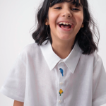 Detalhe da Camisa Tom infantil branca em linho vestida por menina, com bordados coloridos de aves brasileiras entre os botões.