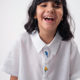 Detalhe da Camisa Tom infantil branca em linho vestida por menina, com bordados coloridos de aves brasileiras entre os botões.