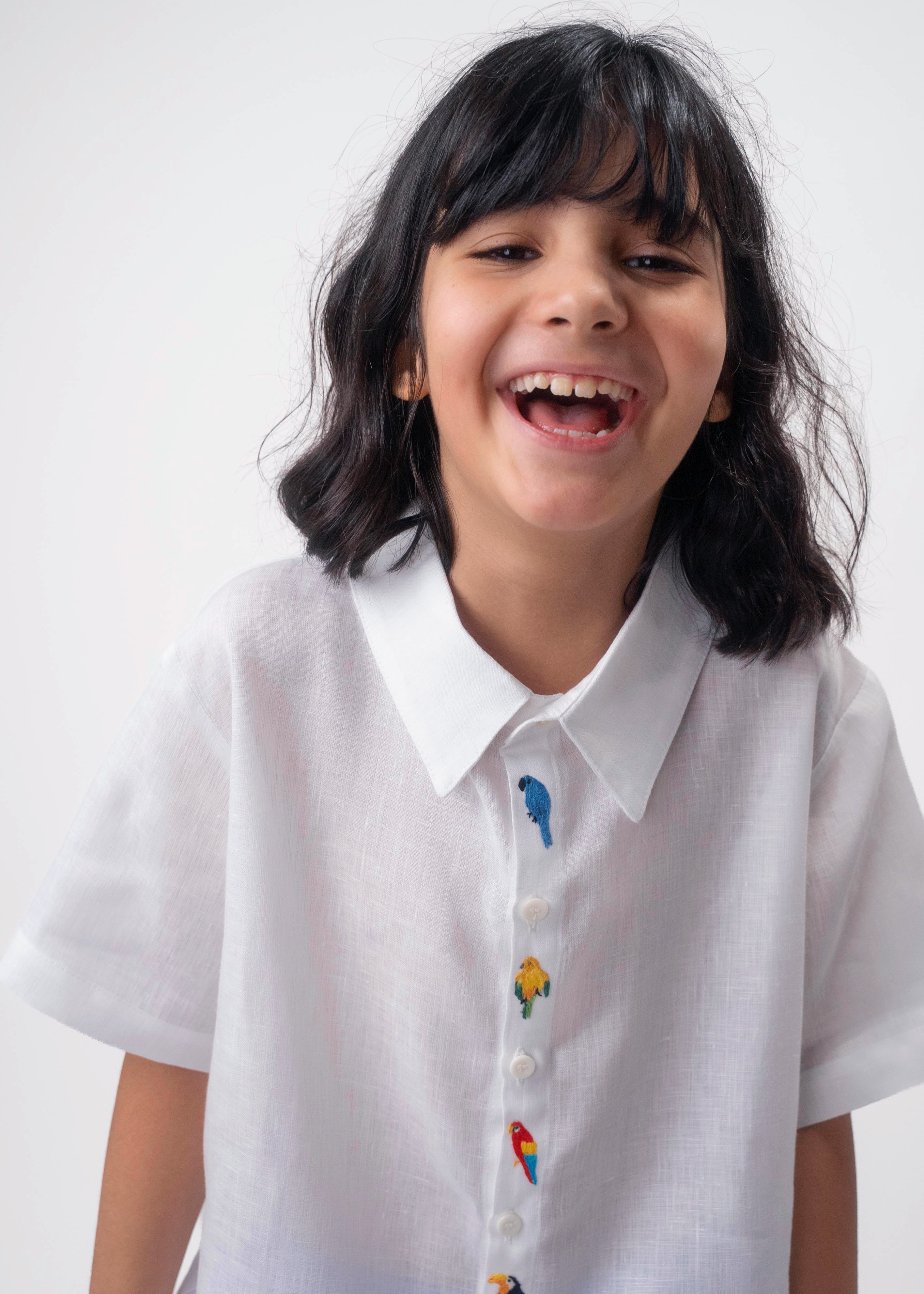 Detalhe da Camisa Tom infantil branca em linho vestida por menina, com bordados coloridos de aves brasileiras entre os botões.