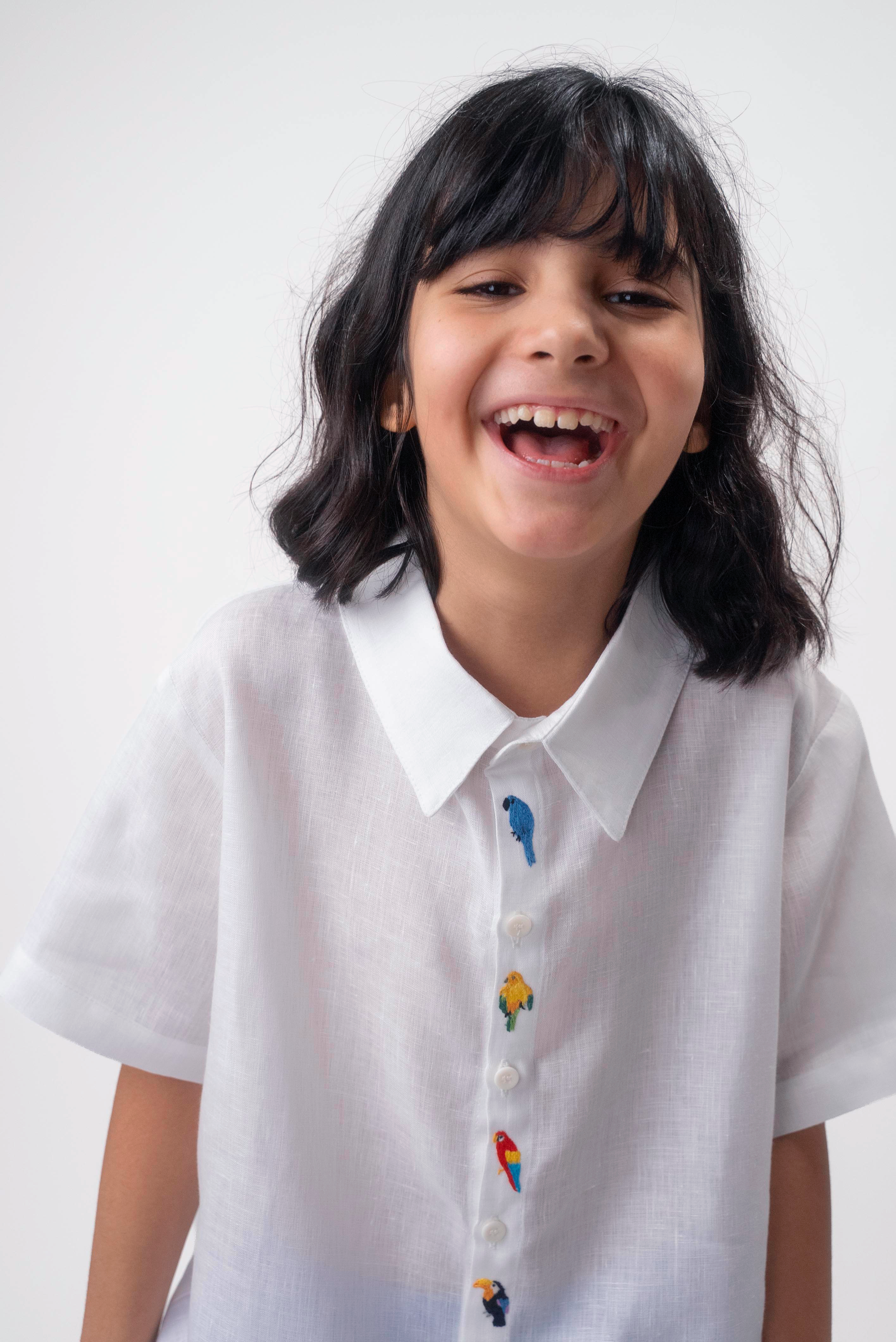 Detalhe da Camisa Tom infantil branca em linho vestida por menina, com bordados coloridos de aves brasileiras entre os botões.