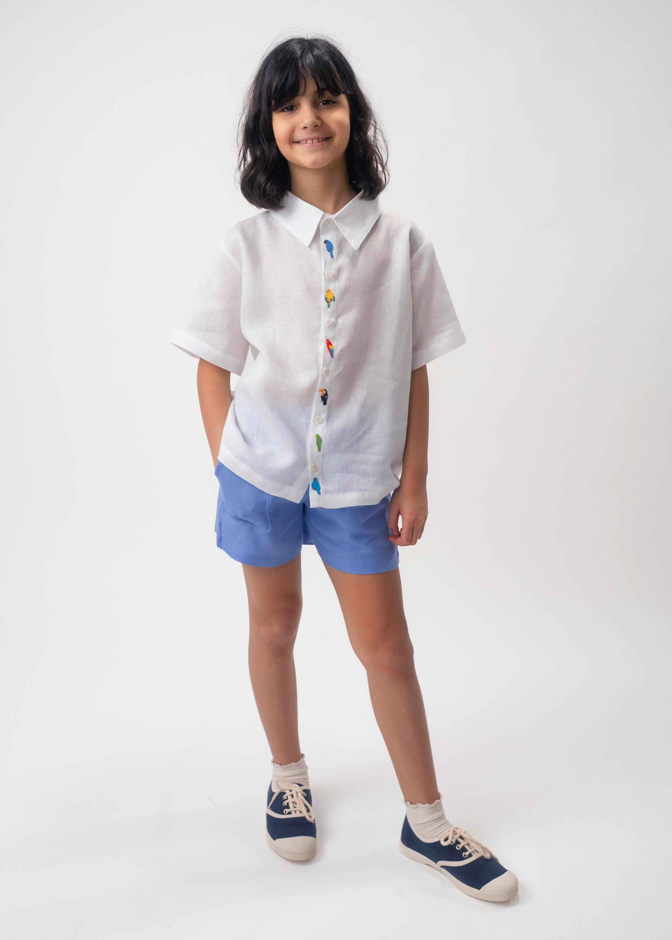 Menina usando Camisa Tom branca em linho com aves brasileiras bordadas à mão e shorts azul.