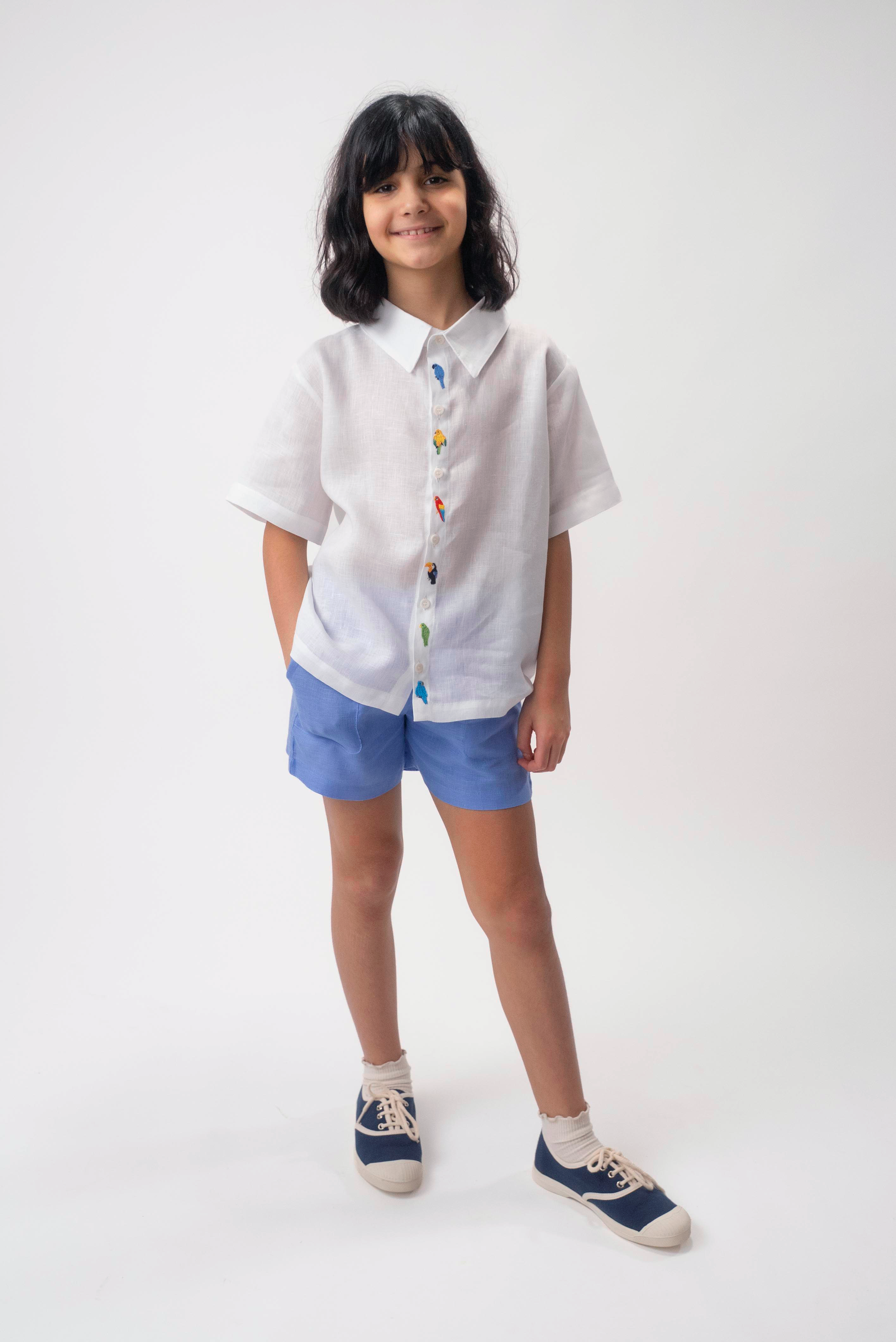 Menina usando Camisa Tom branca em linho com aves brasileiras bordadas à mão e shorts azul.