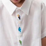 Detalhe da Camisa Tom branca em linho infantil com gola clássica e pássaros coloridos bordados à mão na vista frontal.