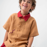 Menino sentado usando Camisa Luca em linho caramelo com gola vermelha contrastante e cobra-coral bordada à mão.