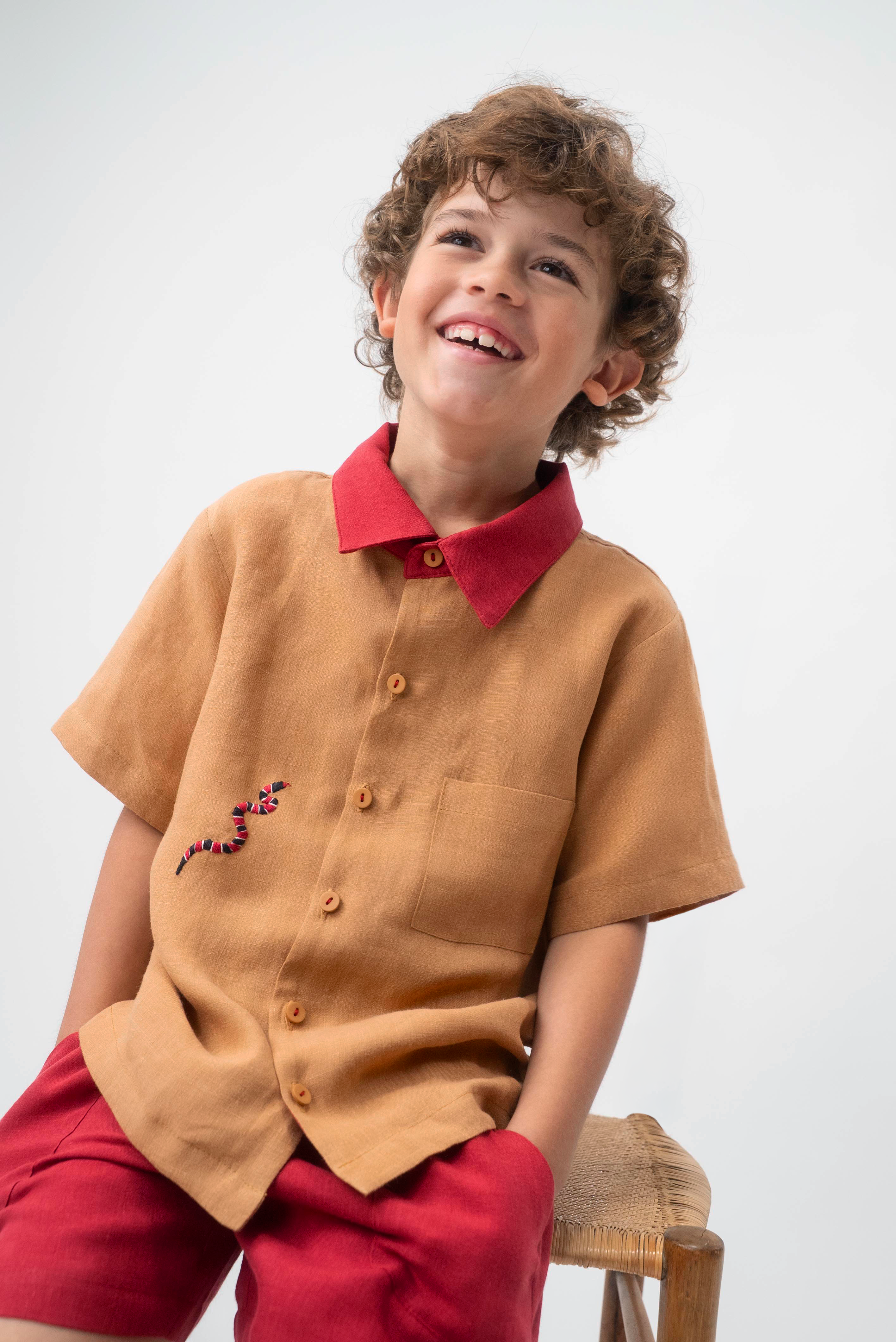 Menino sentado usando Camisa Luca em linho caramelo com gola vermelha contrastante e cobra-coral bordada à mão.