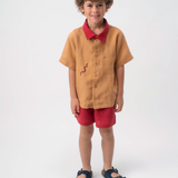 Menino usando Camisa Luca infantil em linho caramelo com gola vermelha e bordado de cobra-coral, combinada com shorts vermelhos.