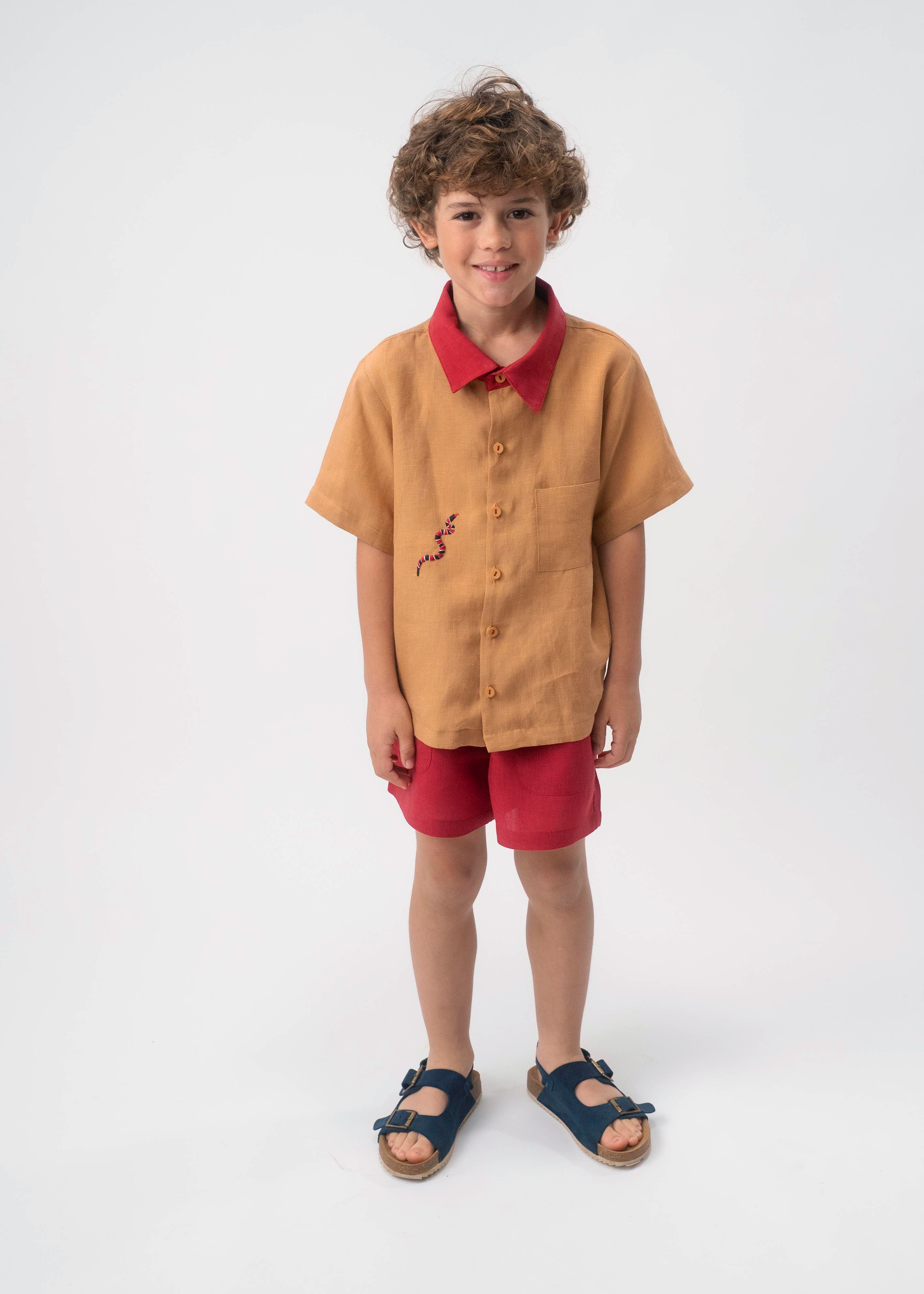 Menino usando Camisa Luca infantil em linho caramelo com gola vermelha e bordado de cobra-coral, combinada com shorts vermelhos.