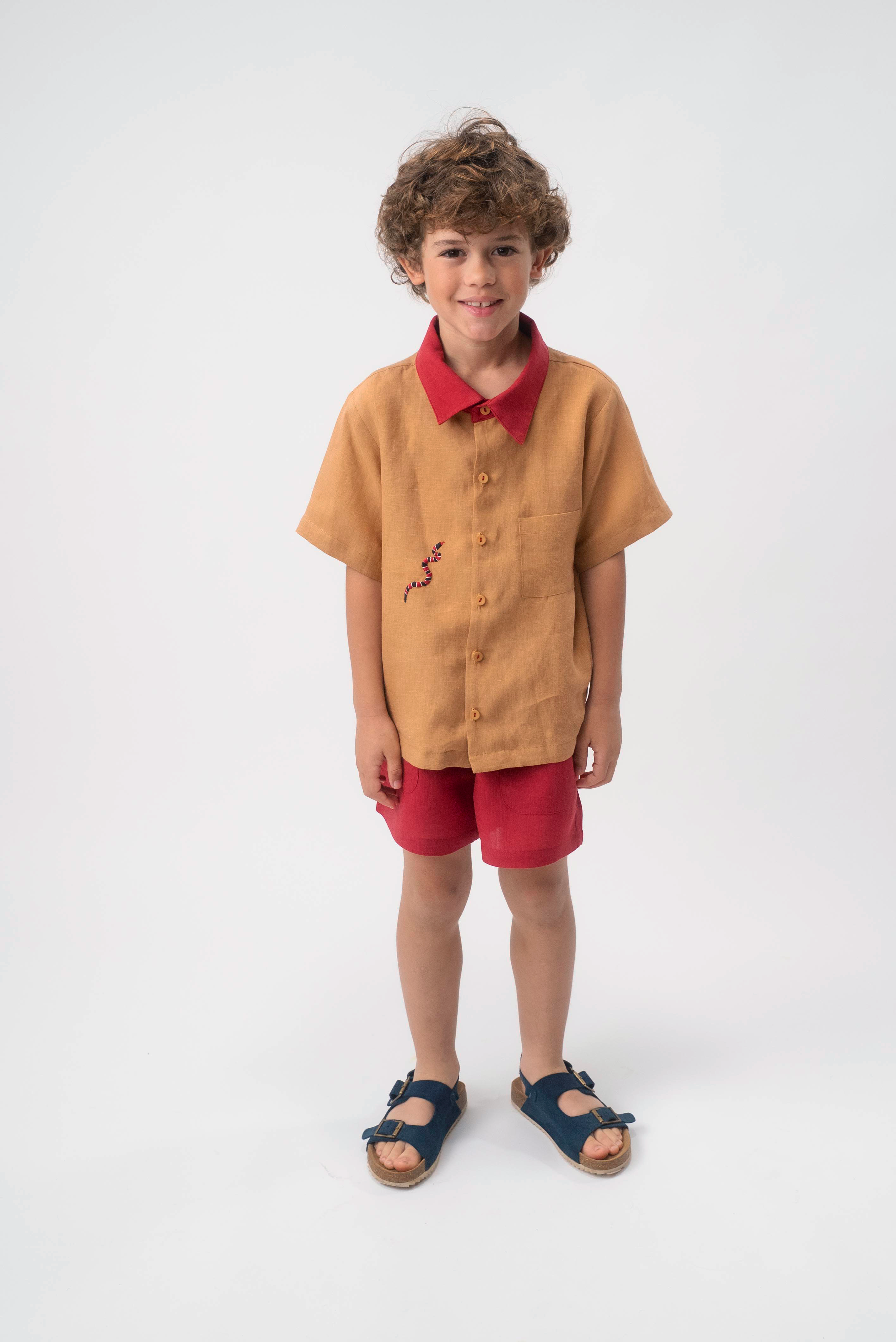 Menino usando Camisa Luca infantil em linho caramelo com gola vermelha e bordado de cobra-coral, combinada com shorts vermelhos.