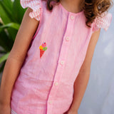 Menina sorrindo usando camisa infantil em linho rosa com mangas de babado listrado e bordado de sorvete de casquinha no peito, em zoom aproximado.