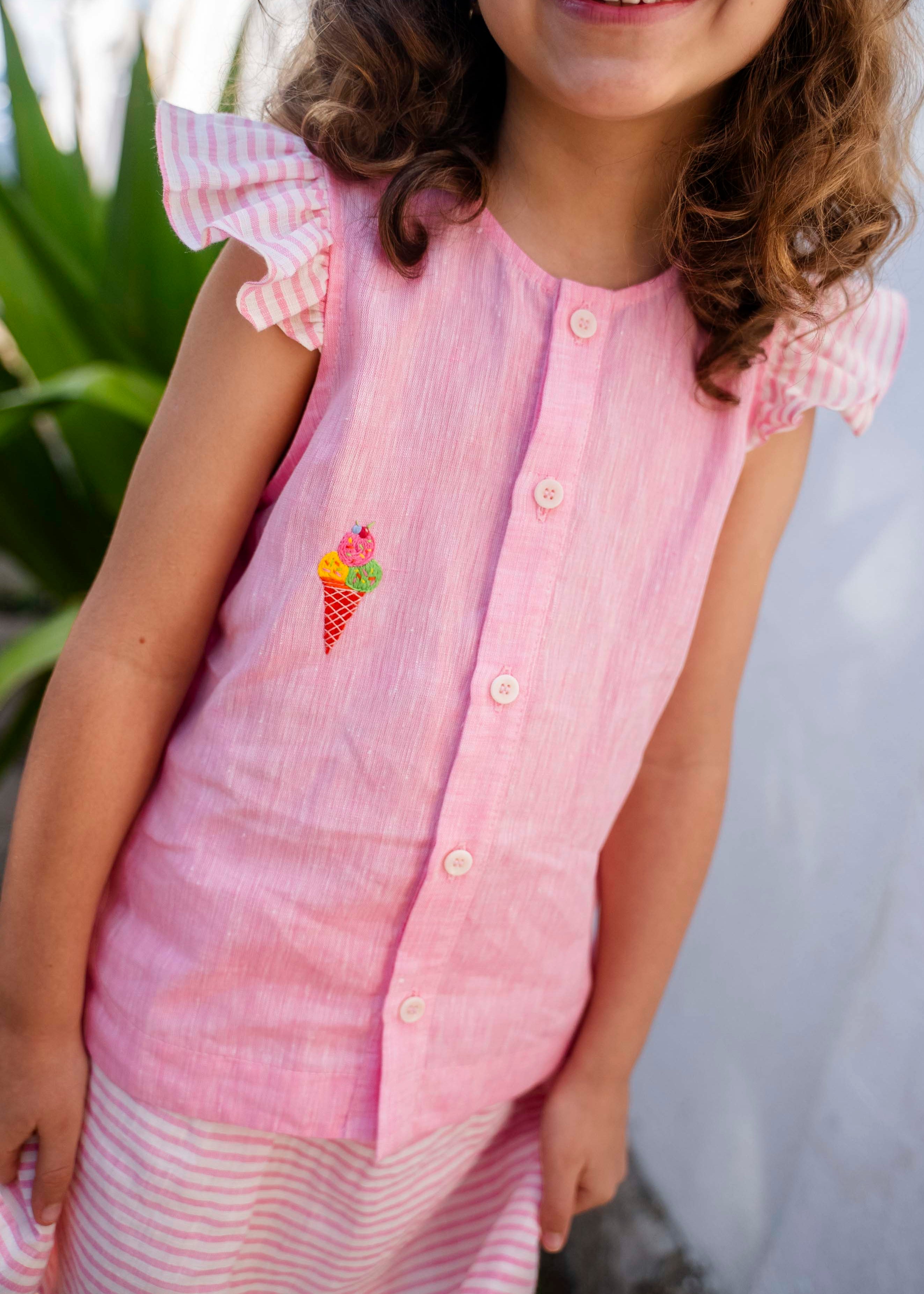 Menina sorrindo usando camisa infantil em linho rosa com mangas de babado listrado e bordado de sorvete de casquinha no peito, em zoom aproximado.