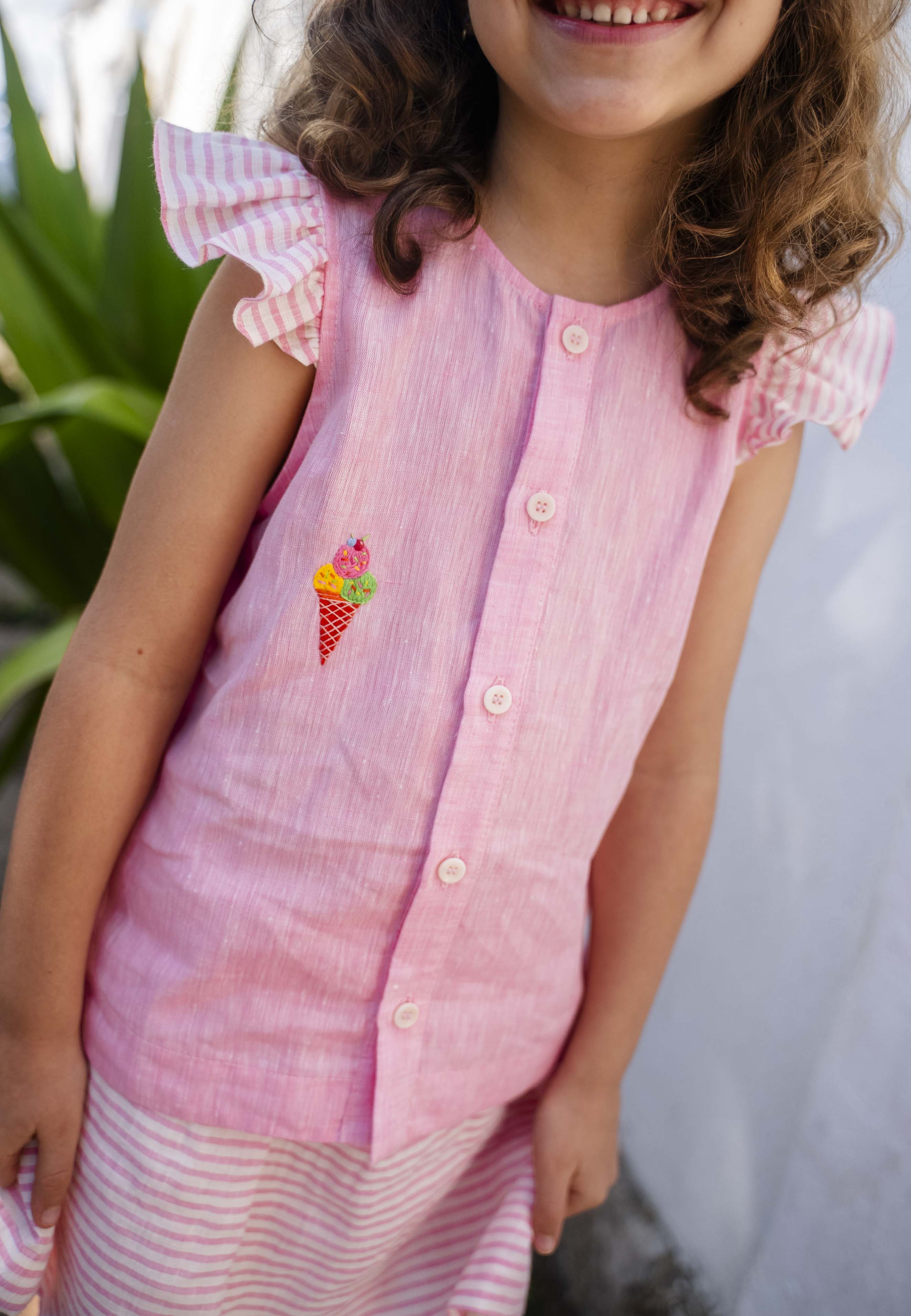 Menina sorrindo usando camisa infantil em linho rosa com mangas de babado listrado e bordado de sorvete de casquinha no peito, em zoom aproximado.