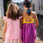 Menina de costas usando camisa infantil em linho rosa com mangas de babado listrado, combinando com uma listrada. Ao lado, amiga de costas com vestido roxo.