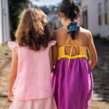 Menina de costas usando camisa infantil em linho rosa com mangas de babado listrado, combinando com uma listrada. Ao lado, amiga de costas com vestido roxo.