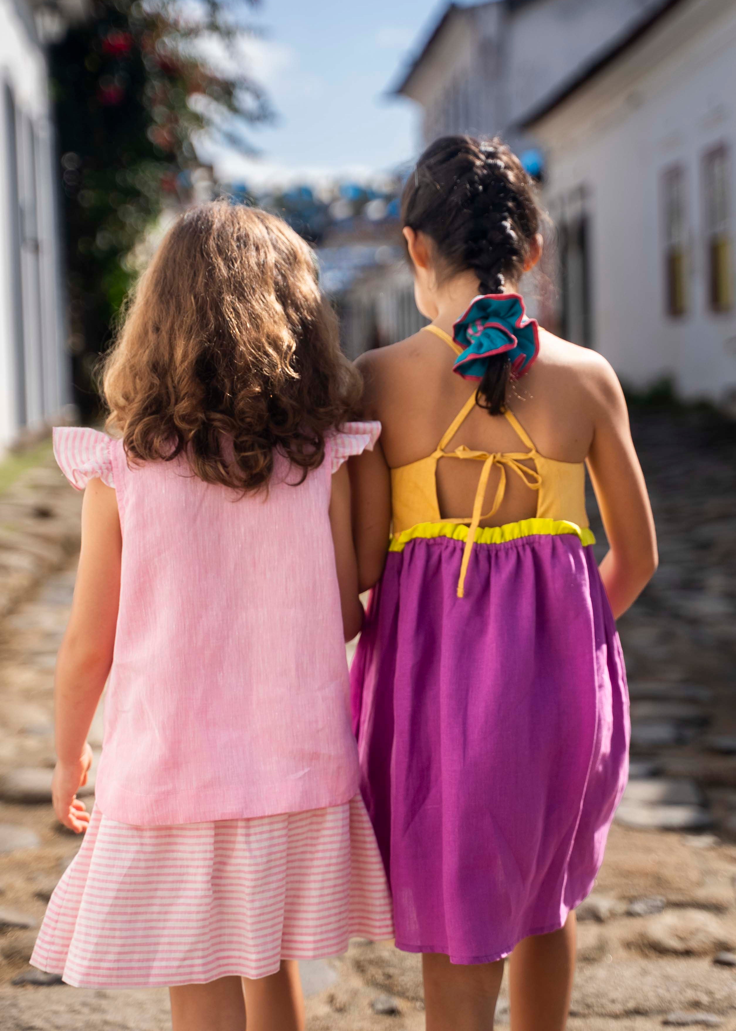 Menina de costas usando camisa infantil em linho rosa com mangas de babado listrado, combinando com uma listrada. Ao lado, amiga de costas com vestido roxo.