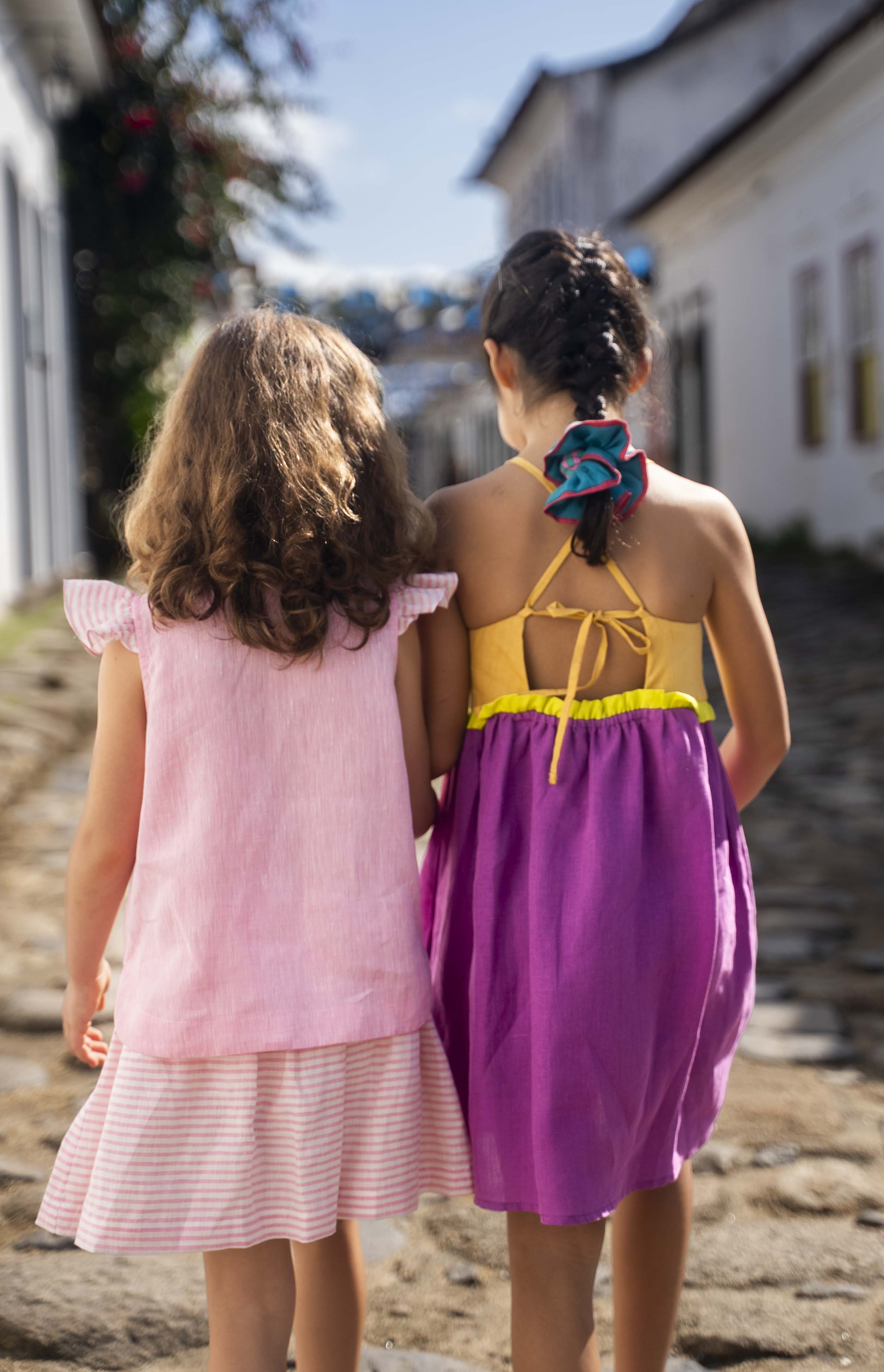 Menina de costas usando camisa infantil em linho rosa com mangas de babado listrado, combinando com uma listrada. Ao lado, amiga de costas com vestido roxo.