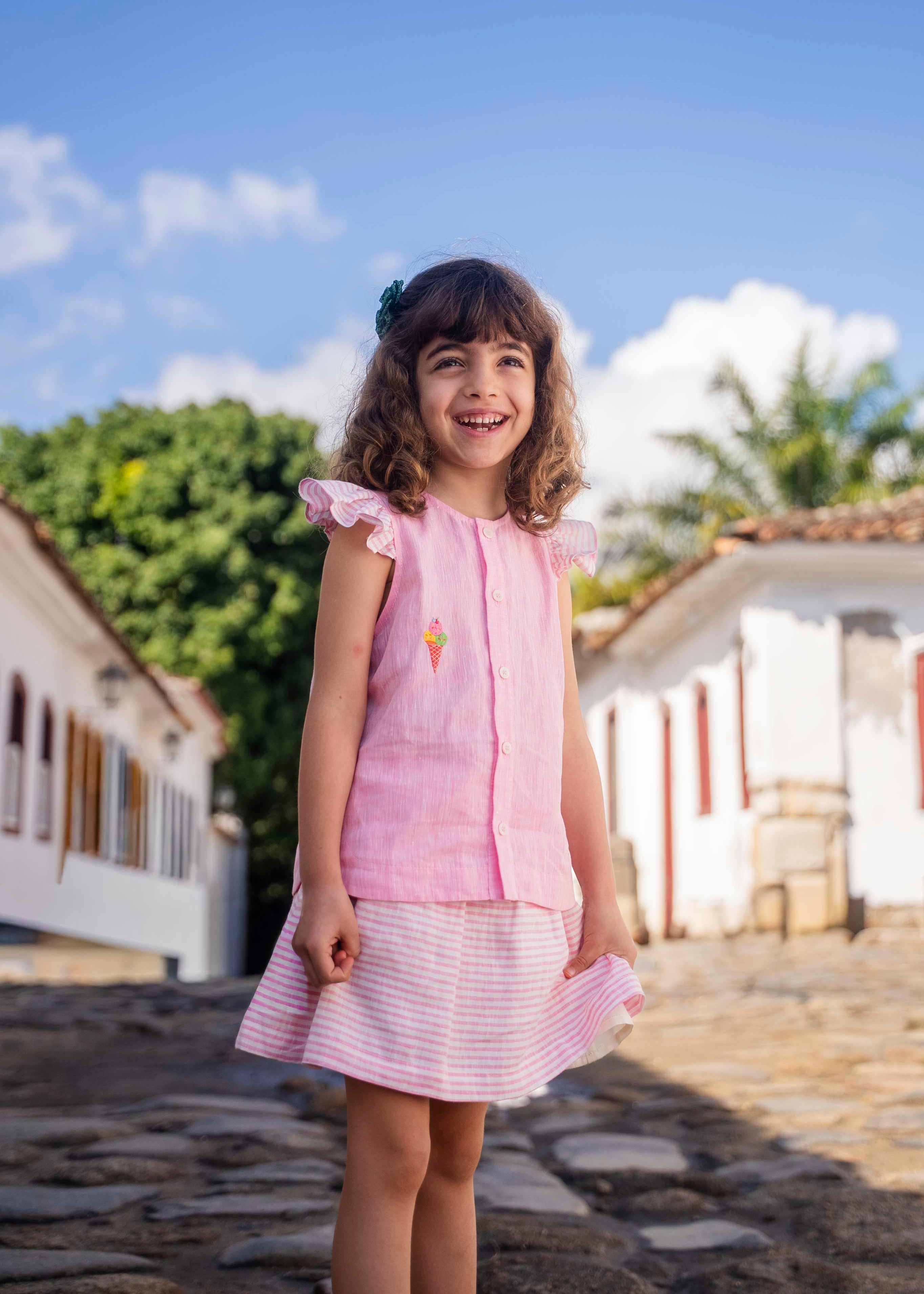 Menina sorrindo usando camisa infantil em linho rosa com mangas de babado listrado e bordado de sorvete de casquinha no peito, combinando com uma saia listrada.