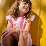 Menina sentada e sorrindo, usando camisa infantil em linho rosa com mangas de babado listrado, combinando com saia listrada.