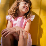 Menina sentada e sorrindo, usando camisa infantil em linho rosa com mangas de babado listrado, combinando com saia listrada.
