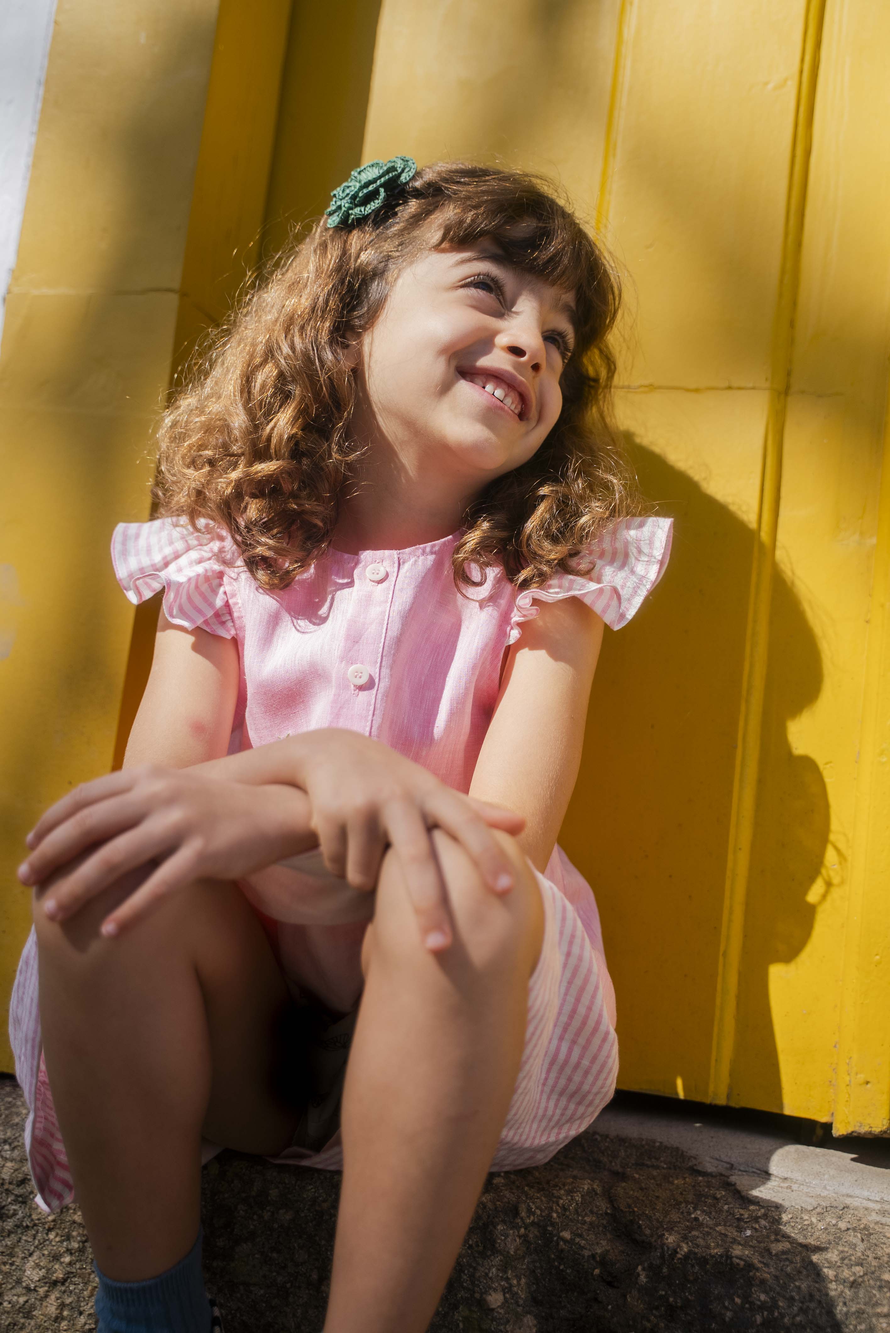 Menina sentada e sorrindo, usando camisa infantil em linho rosa com mangas de babado listrado, combinando com saia listrada.