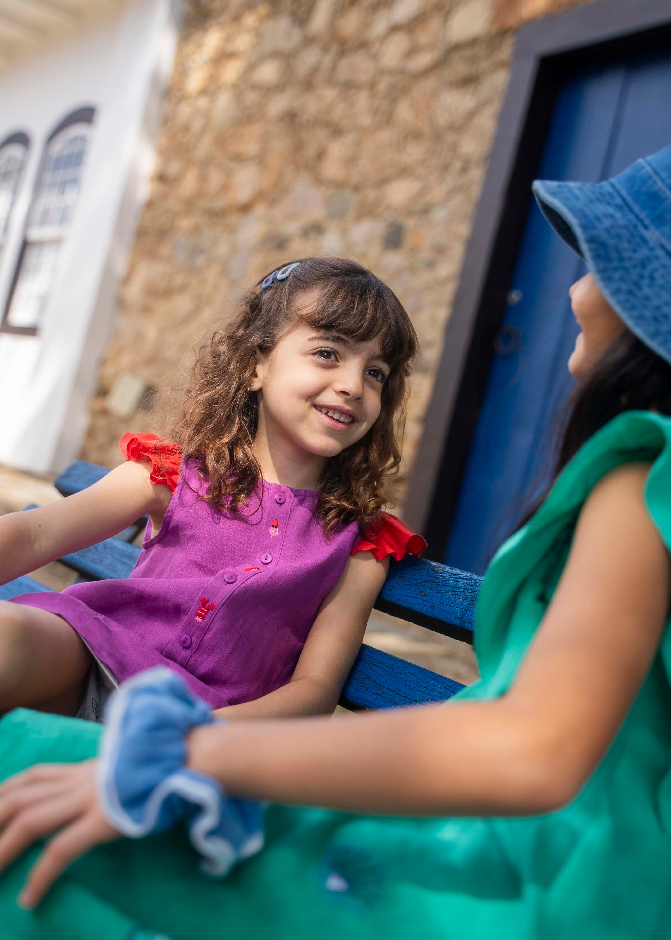 Menina usando camisa infantil em linho roxo com mangas com babado vermelho e bordado de bichinhos do fundo do mar entre os botões.
