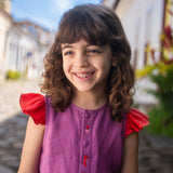 Menina sorrindo usando camisa infantil em linho roxo com mangas de babado vermelho e bordado de bichinhos do fundo do mar entre os botões.