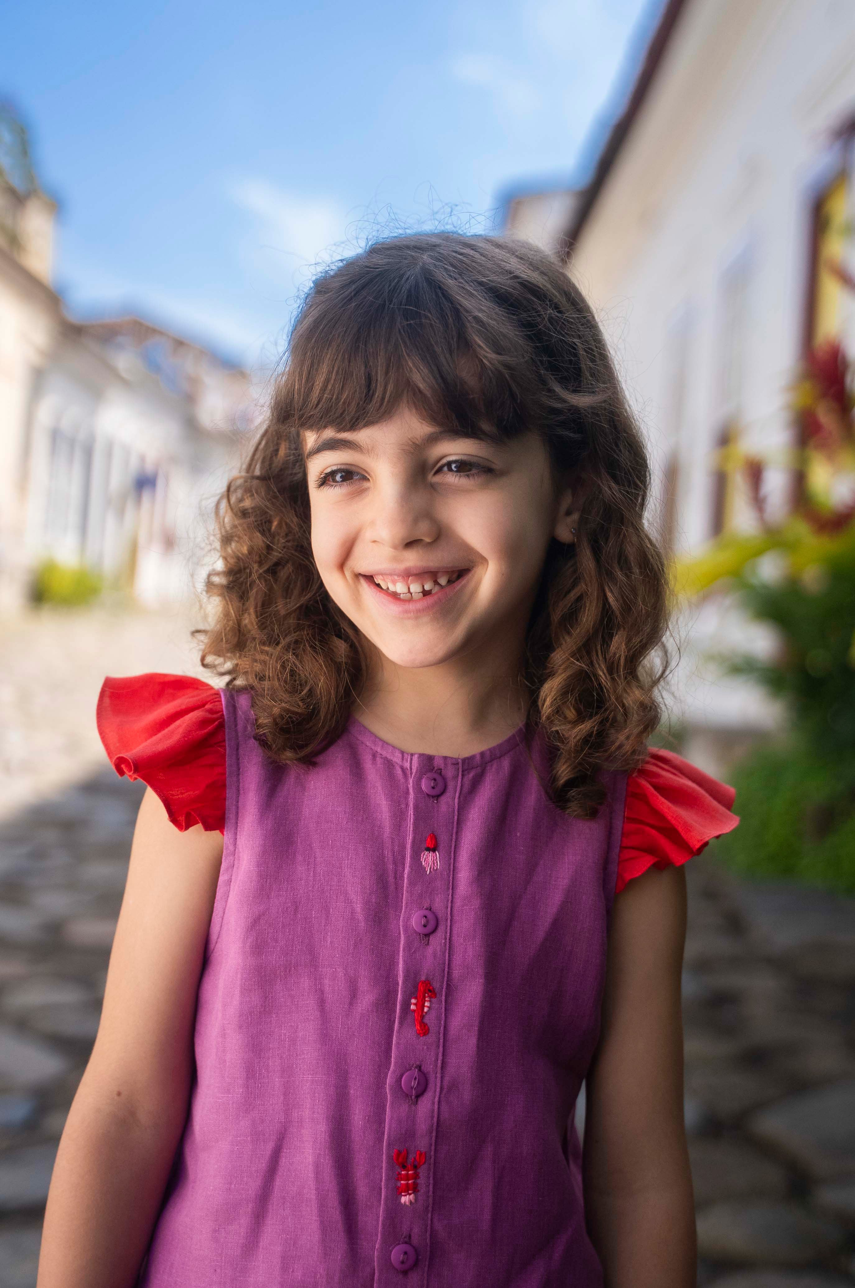 Menina sorrindo usando camisa infantil em linho roxo com mangas de babado vermelho e bordado de bichinhos do fundo do mar entre os botões.