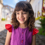 Menina sorrindo usando camisa infantil em linho roxo com mangas de babado vermelho e bordado de bichinhos do fundo do mar entre os botões.