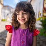 Menina sorrindo usando camisa infantil em linho roxo com mangas de babado vermelho e bordado de bichinhos do fundo do mar entre os botões.