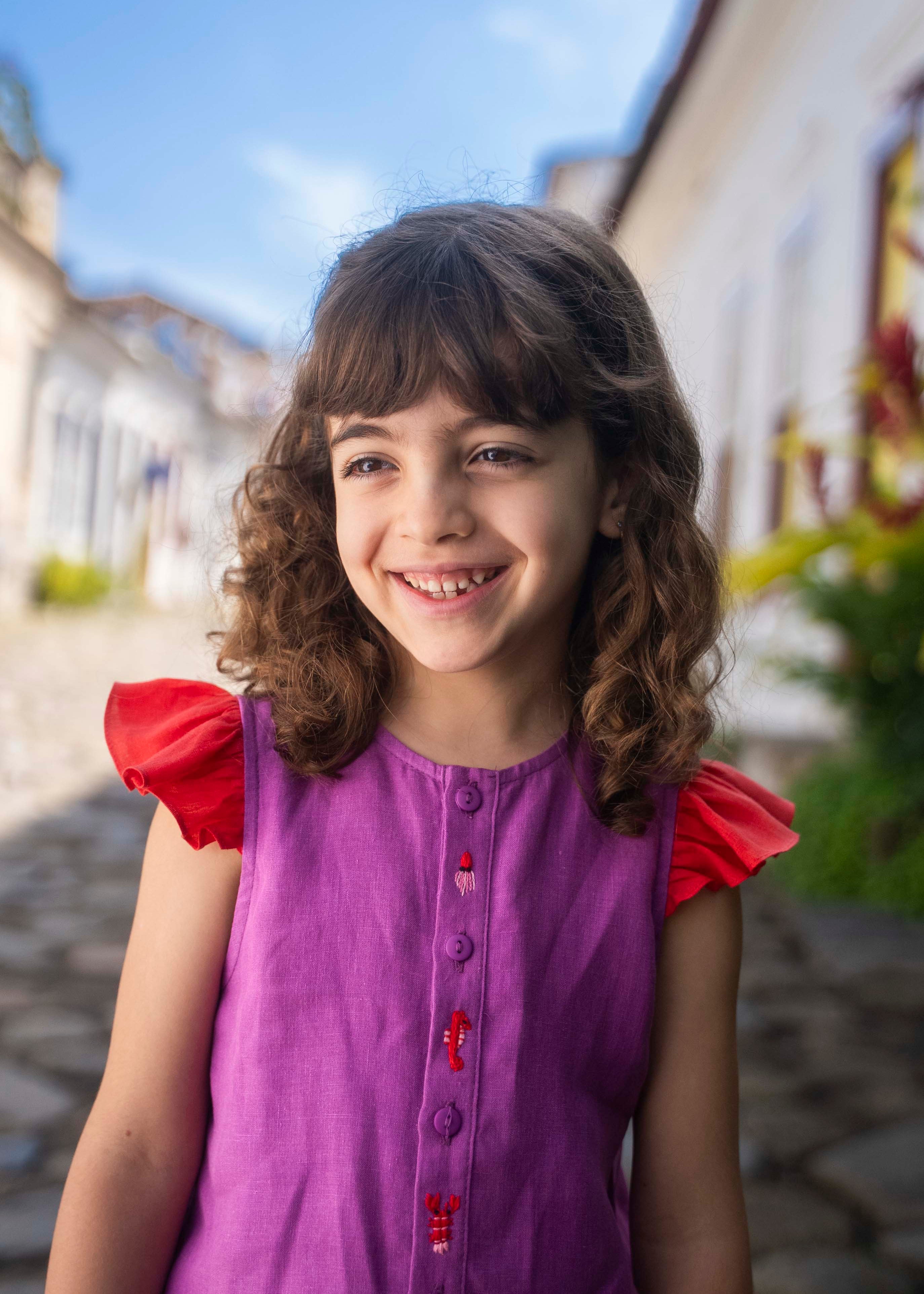 Menina sorrindo usando camisa infantil em linho roxo com mangas de babado vermelho e bordado de bichinhos do fundo do mar entre os botões.