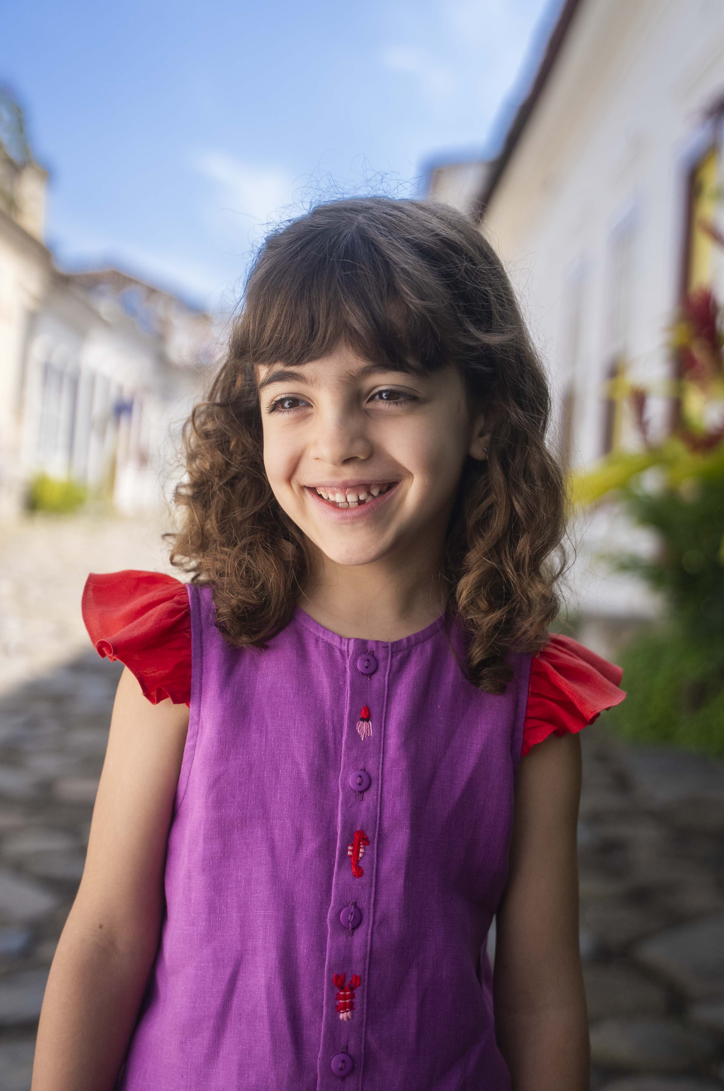 Menina sorrindo usando camisa infantil em linho roxo com mangas de babado vermelho e bordado de bichinhos do fundo do mar entre os botões.