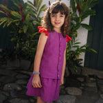 Menina usando camisa infantil em linho roxo com mangas de babado vermelho e bordado de bichinhos do fundo do mar entre os botões, com saia combinando.