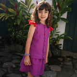 Menina usando camisa infantil em linho roxo com mangas de babado vermelho e bordado de bichinhos do fundo do mar entre os botões, com saia combinando.