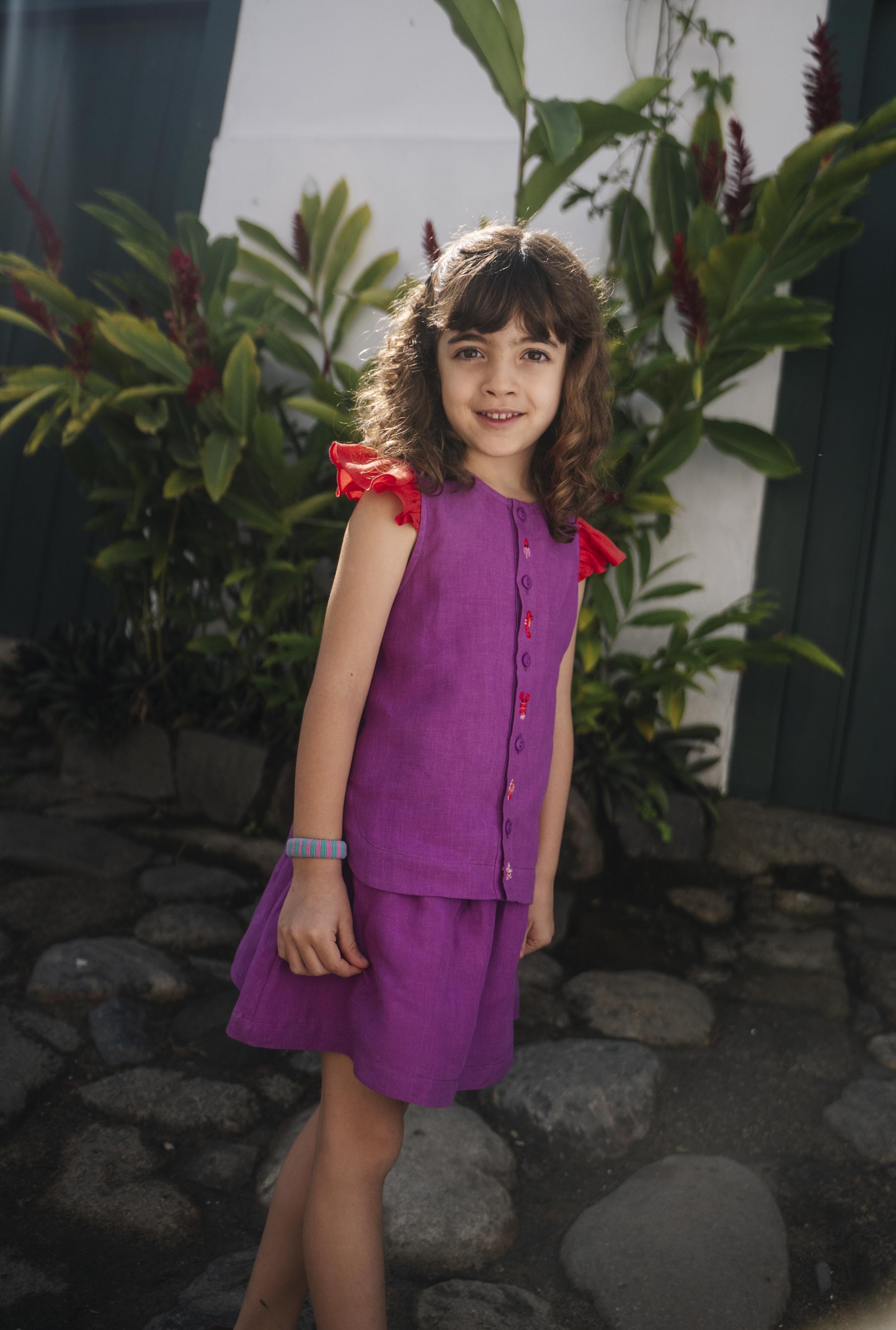 Menina usando camisa infantil em linho roxo com mangas de babado vermelho e bordado de bichinhos do fundo do mar entre os botões, com saia combinando.
