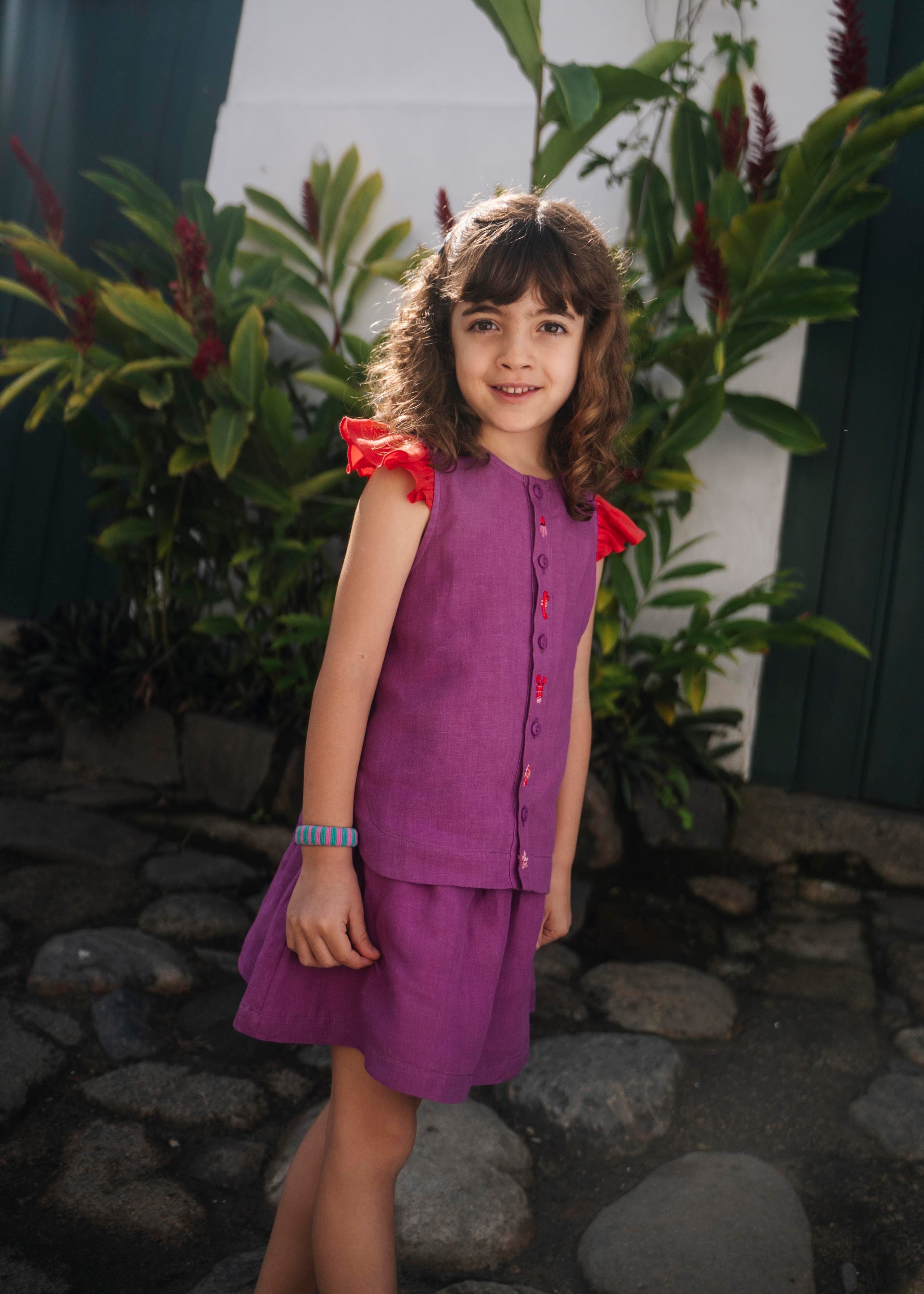 Menina usando camisa infantil em linho roxo com mangas de babado vermelho e bordado de bichinhos do fundo do mar entre os botões, com saia combinando.