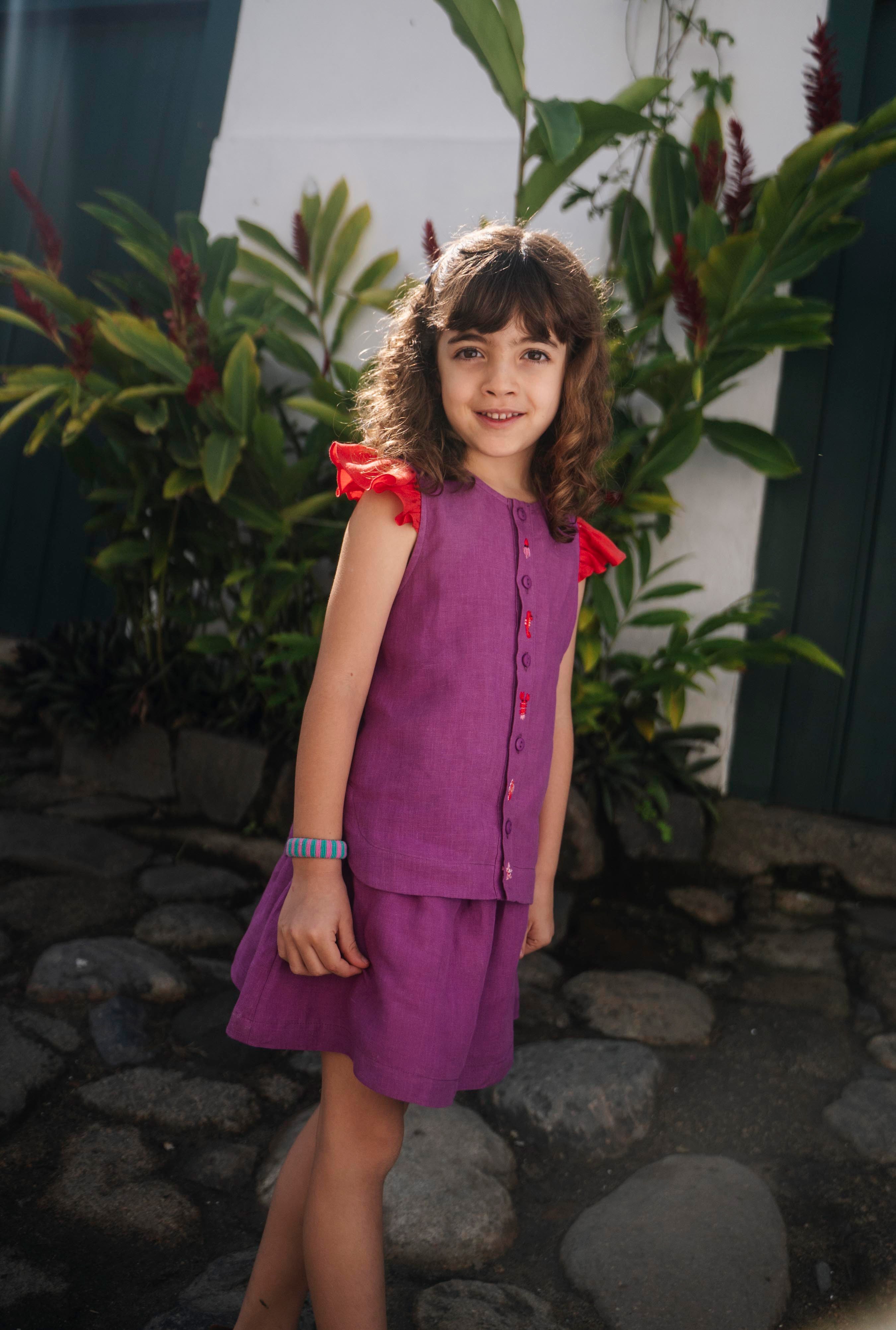 Menina usando camisa infantil em linho roxo com mangas de babado vermelho e bordado de bichinhos do fundo do mar entre os botões, com saia combinando.