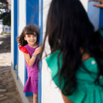 Menina brincando de esconder, vestindo camisa infantil em linho roxo com mangas de babado vermelho, combinando com saia de mesmo tom.