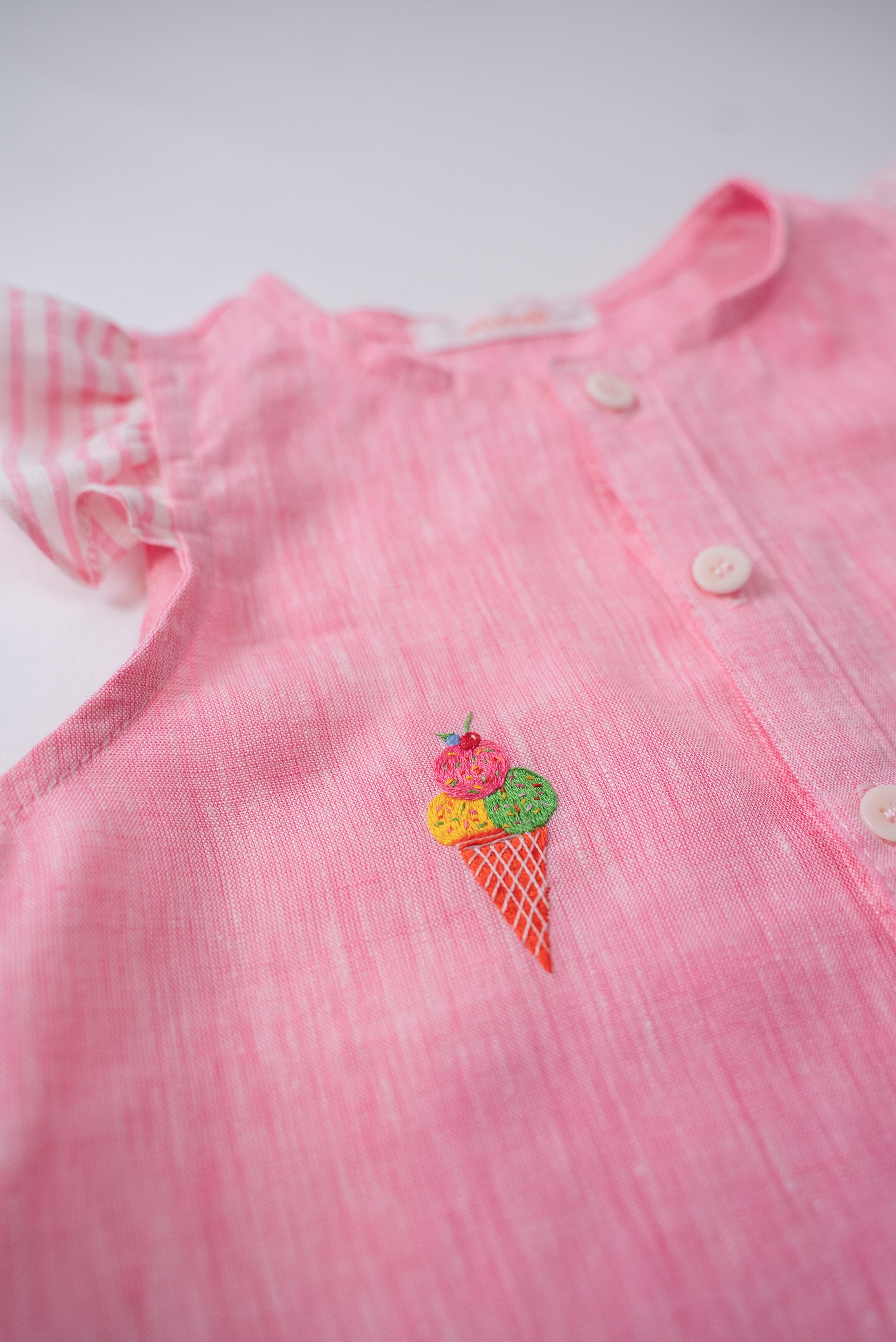 Detalhe do bordado à mão de sorvete de casquinha no peito da camisa infantil menina em linho rosa com mangas de babado listrado.